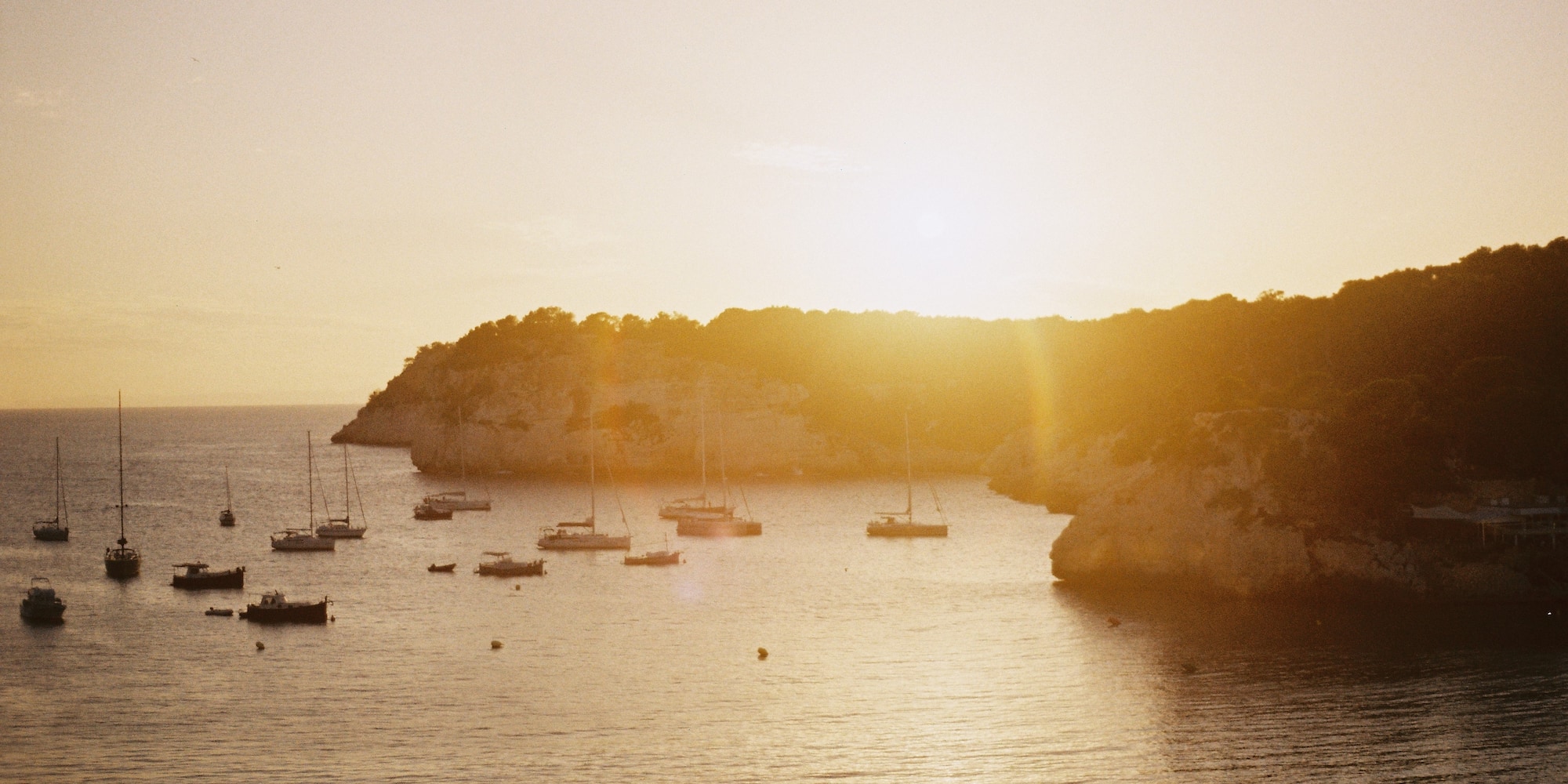 Atardecer dorado en cala con barcos, reflejos en el mar y costa rocosa.