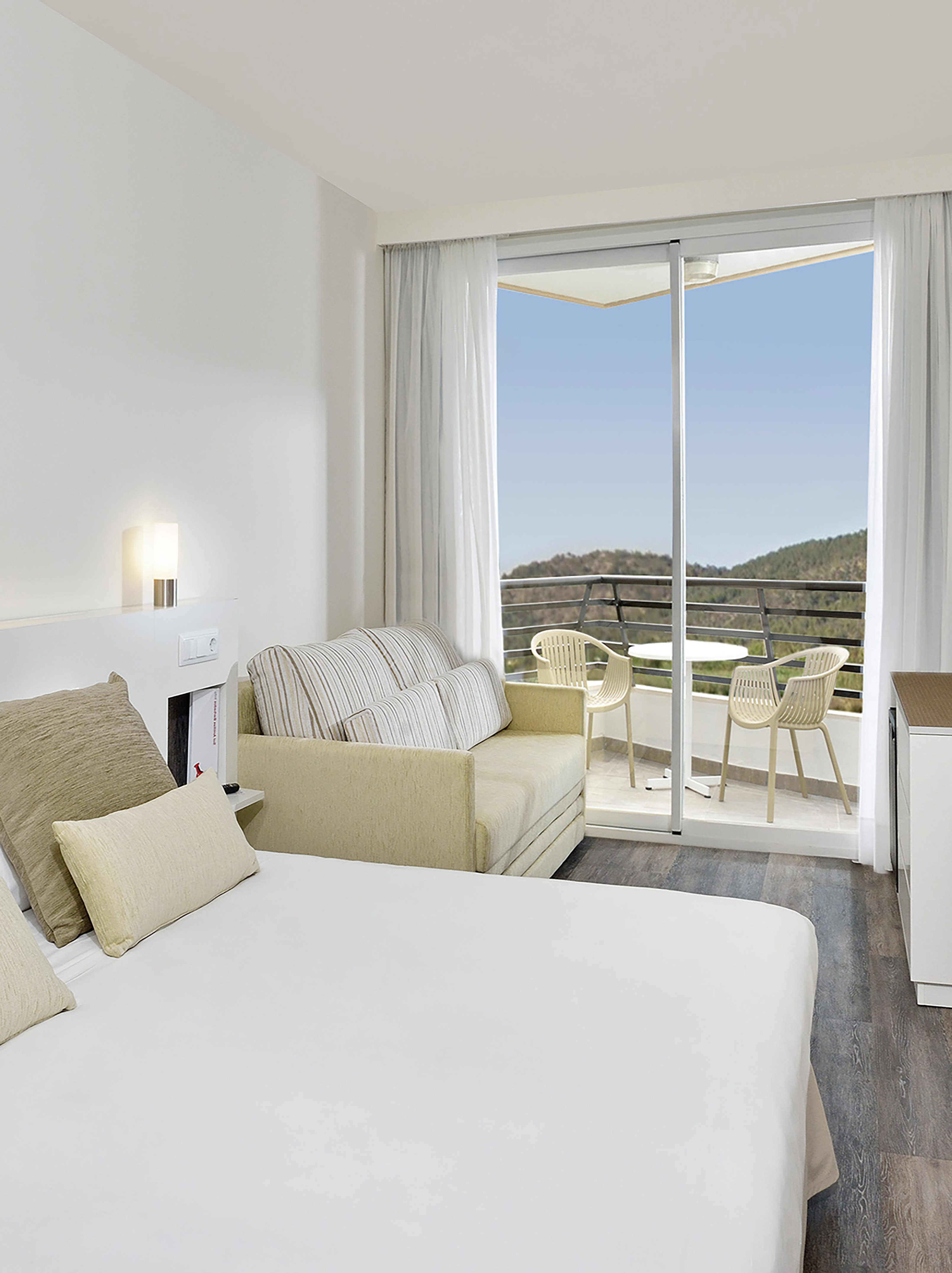 Modern hotel bedroom with white bed, sofa, and balcony overlooking mountains.