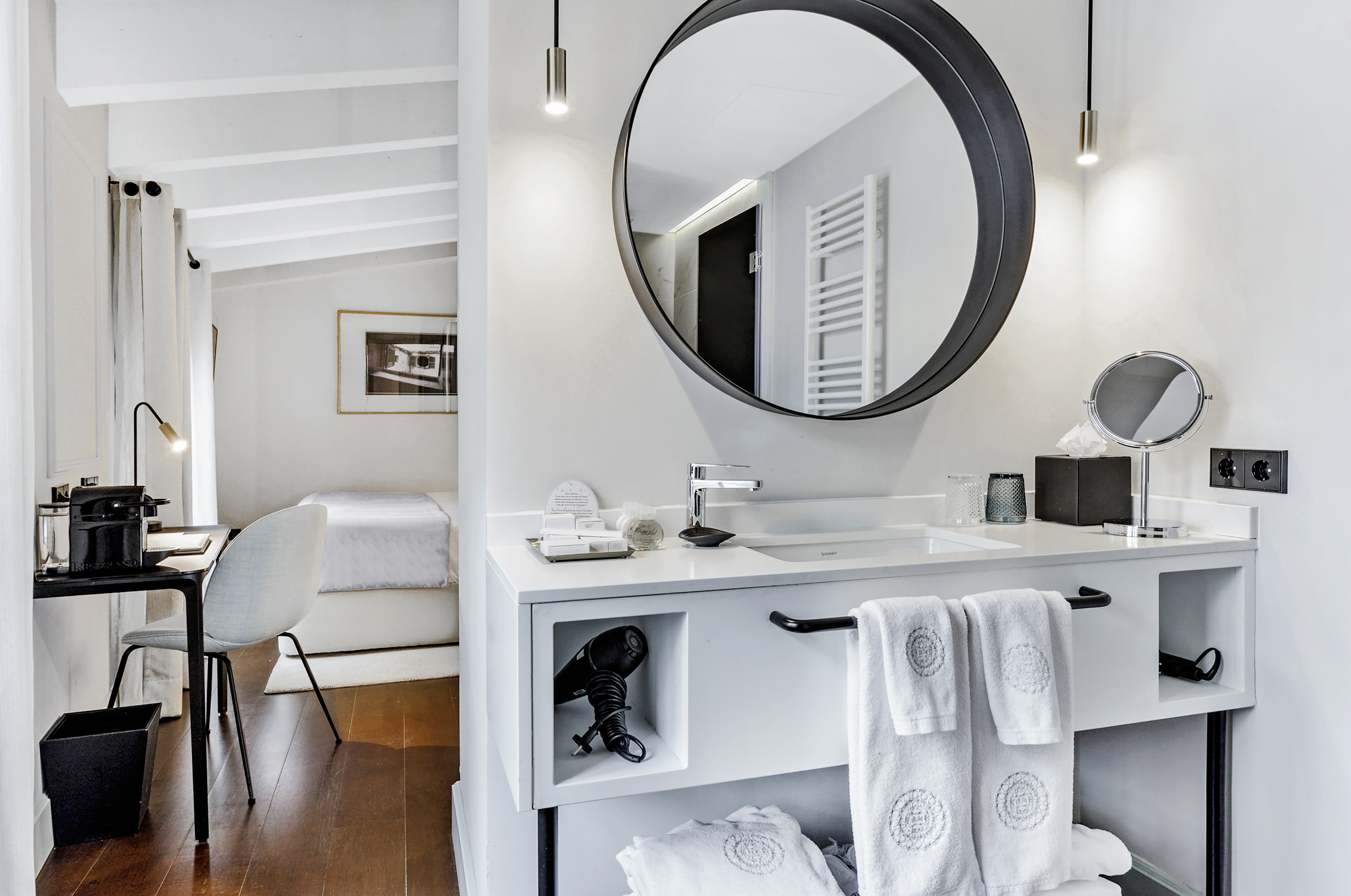 Modern hotel bathroom vanity with round mirror, sink, and bedroom view.