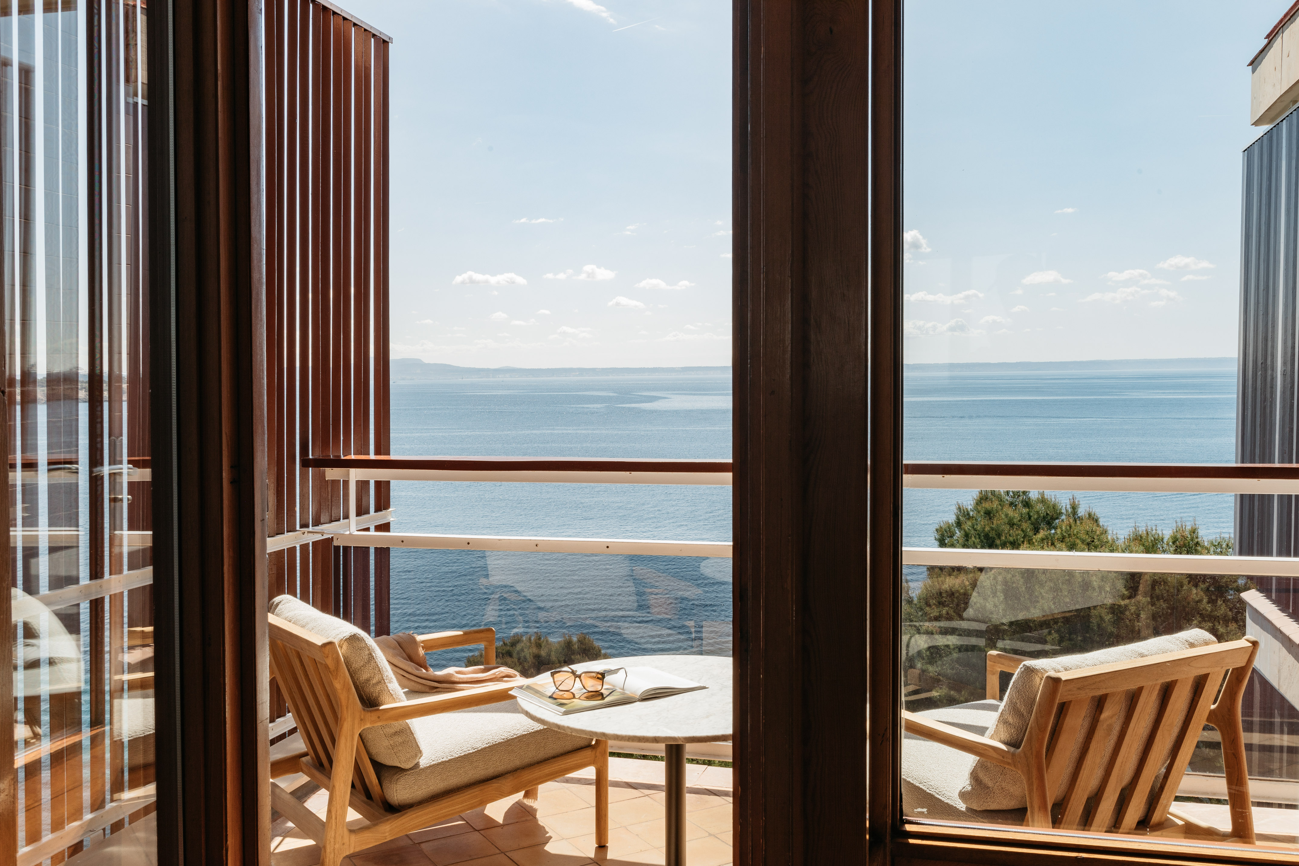 Hotel balcony with wooden chairs, table, and panoramic ocean view.