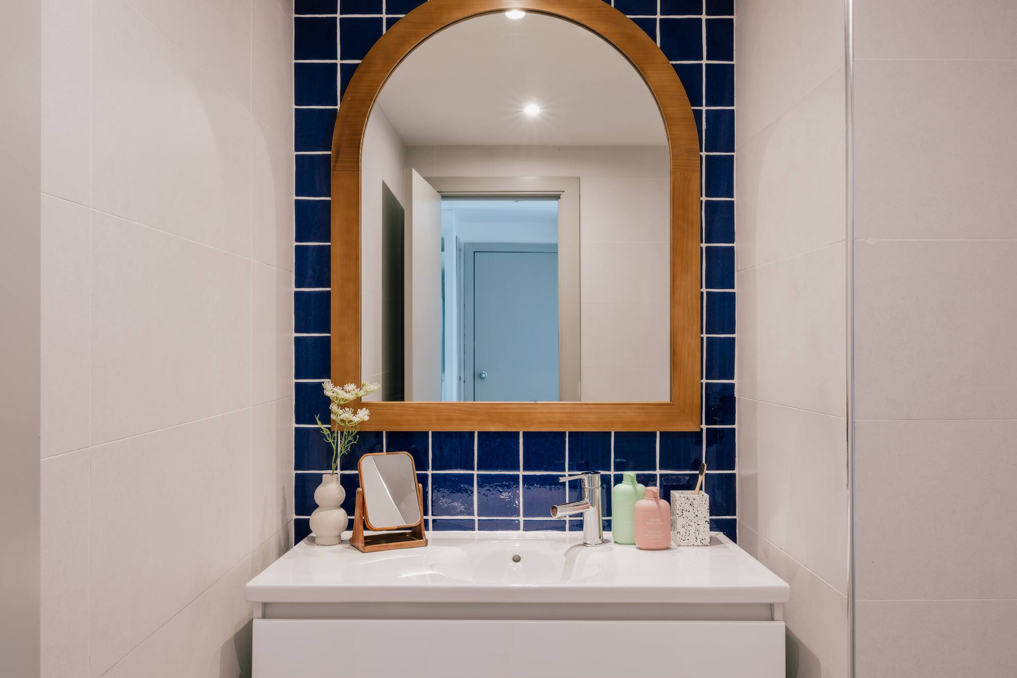White bathroom vanity with arched wooden mirror and blue square tile backsplash.