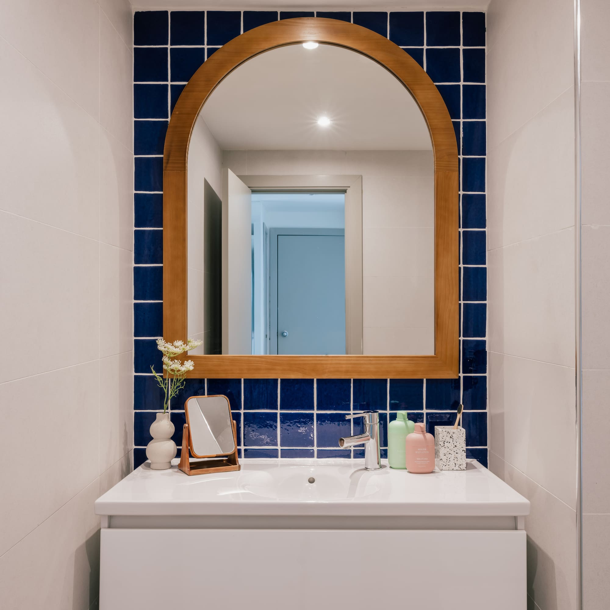 White bathroom vanity with arched wooden mirror and blue square tile backsplash.