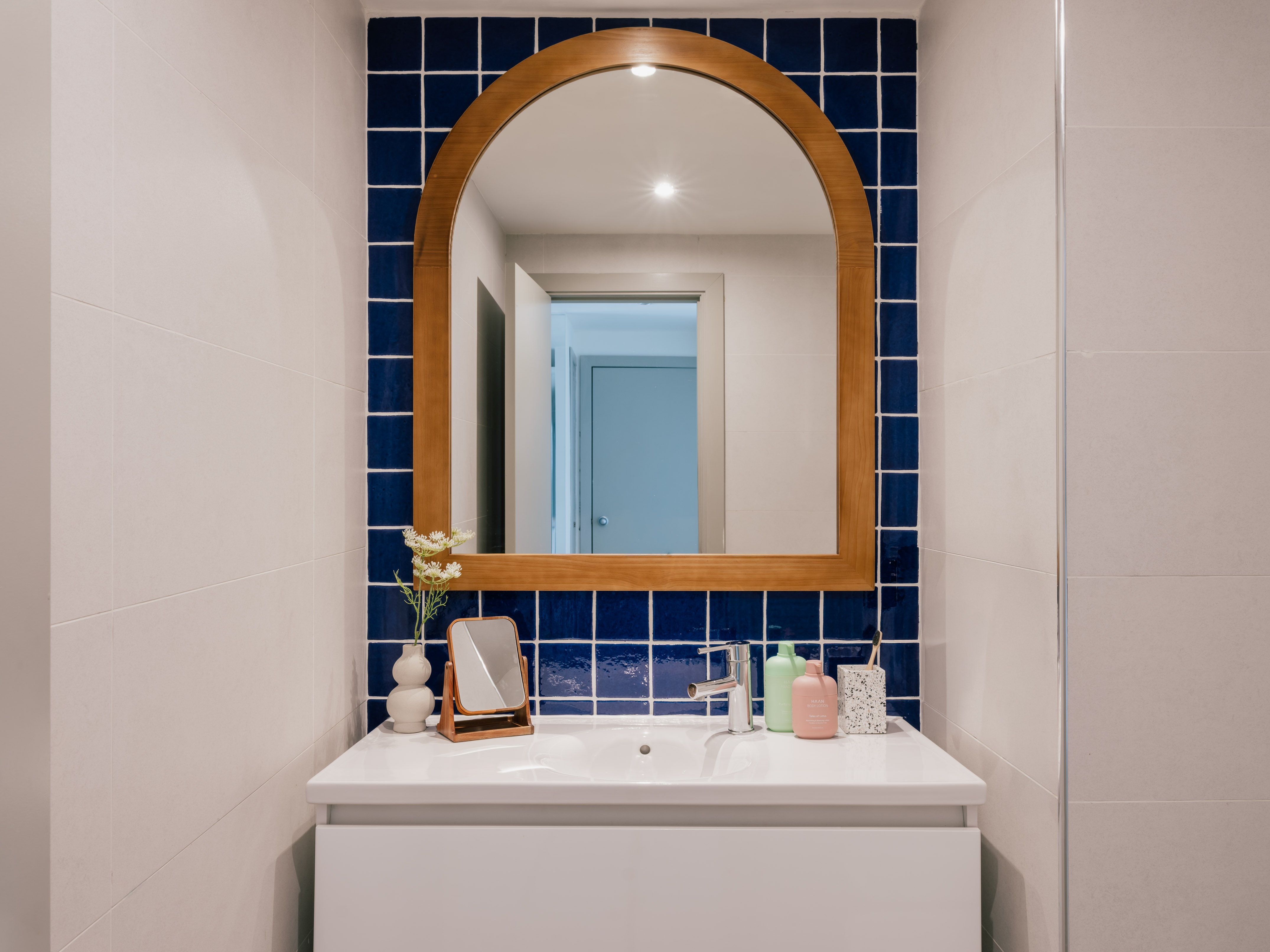 White bathroom vanity with arched wooden mirror and blue square tile backsplash.