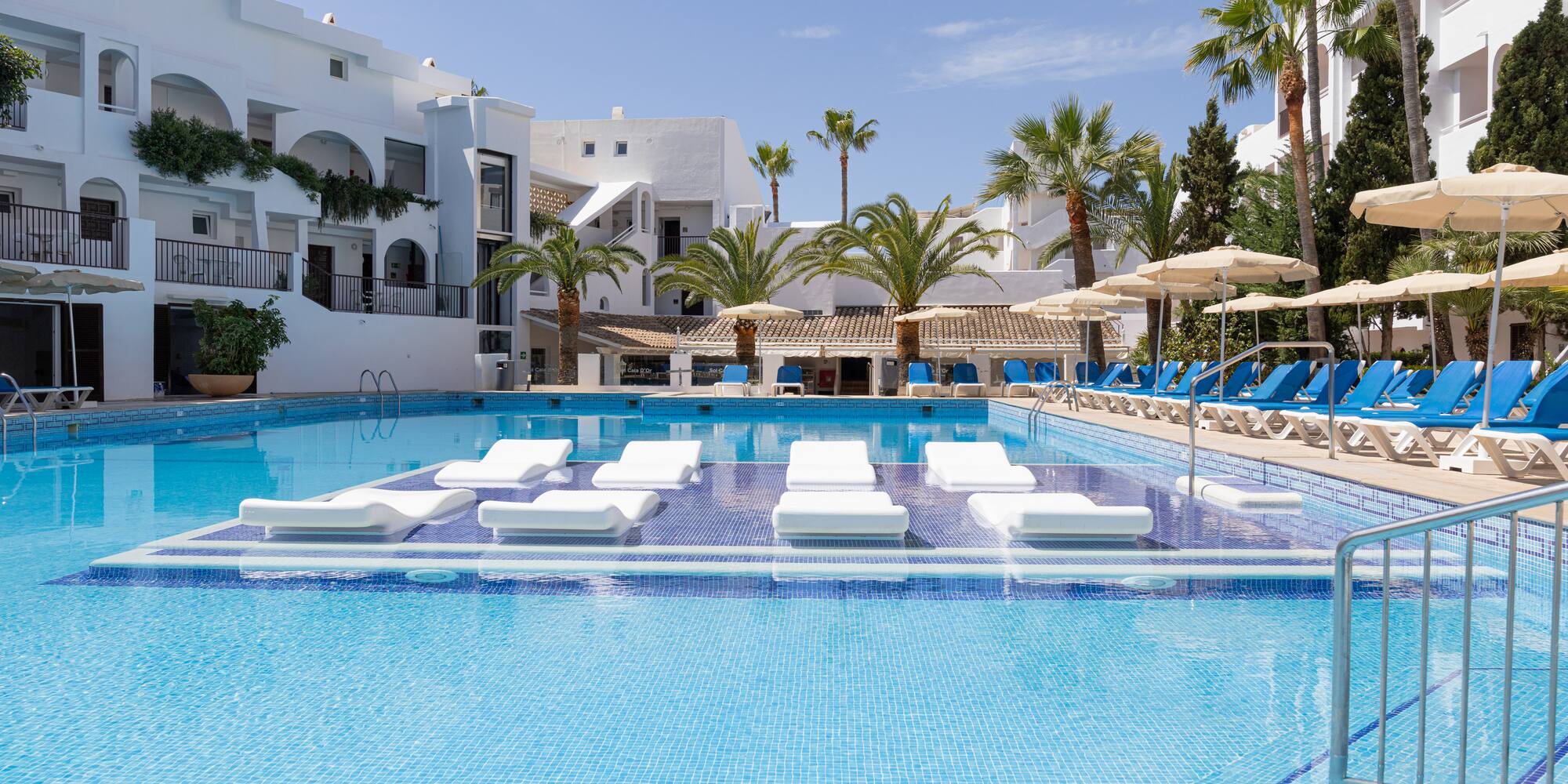 Sunny resort pool with white buildings, palm trees, and submerged loungers.