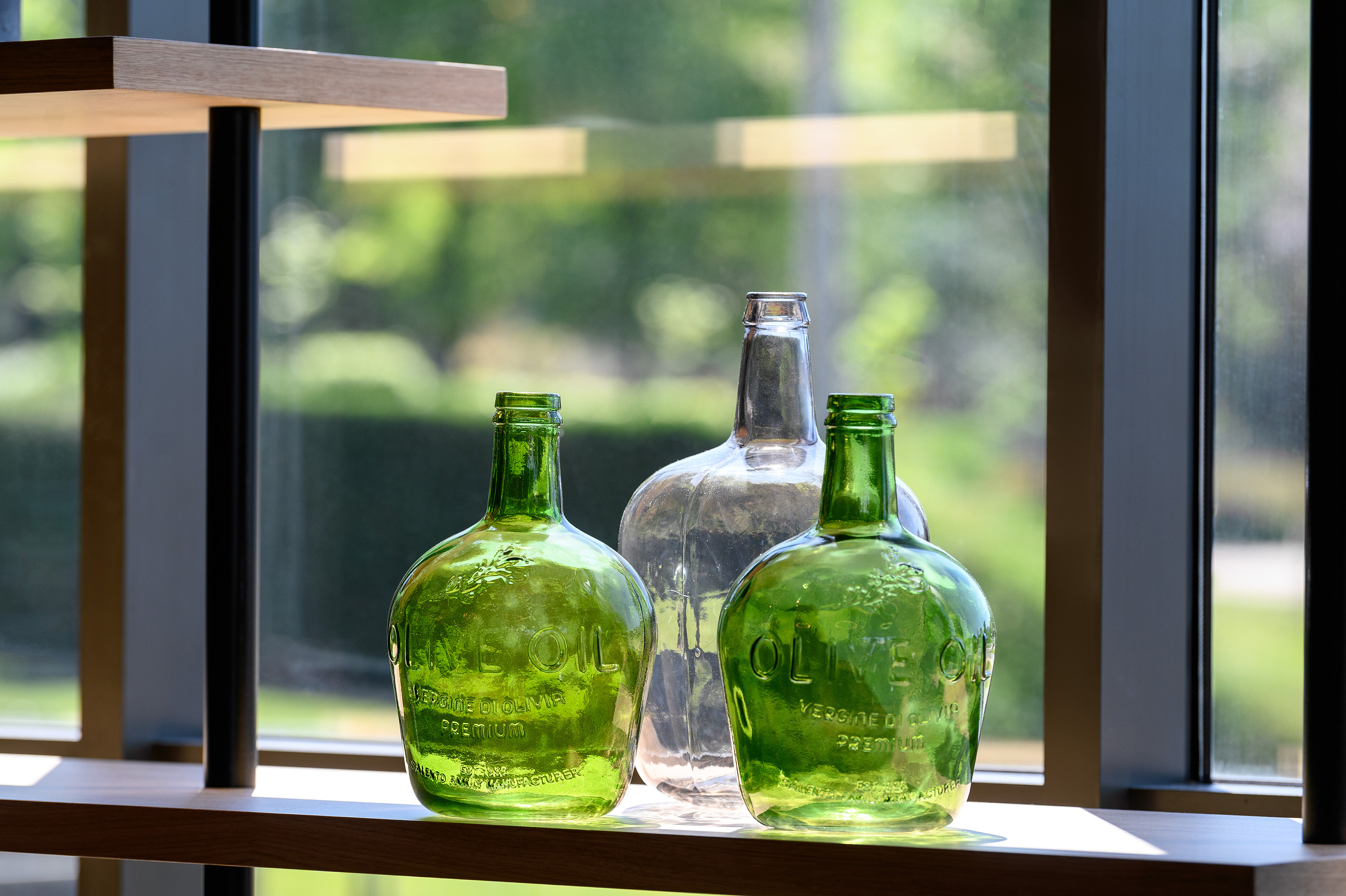 Embossed green and clear olive oil bottles on a wooden shelf by a window.