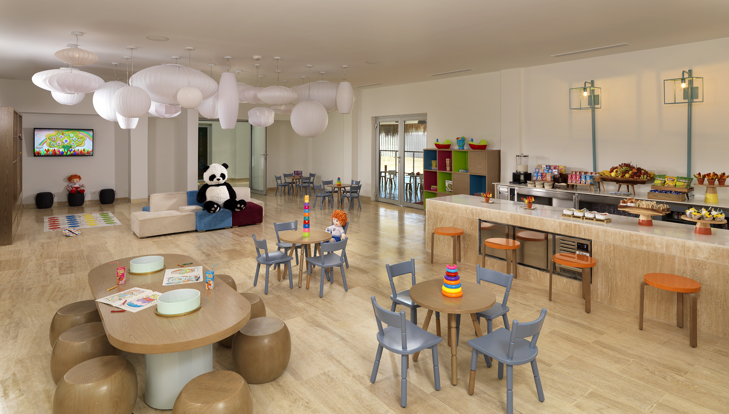 Bright hotel kids club with play tables, sofa, TV, and snack bar.