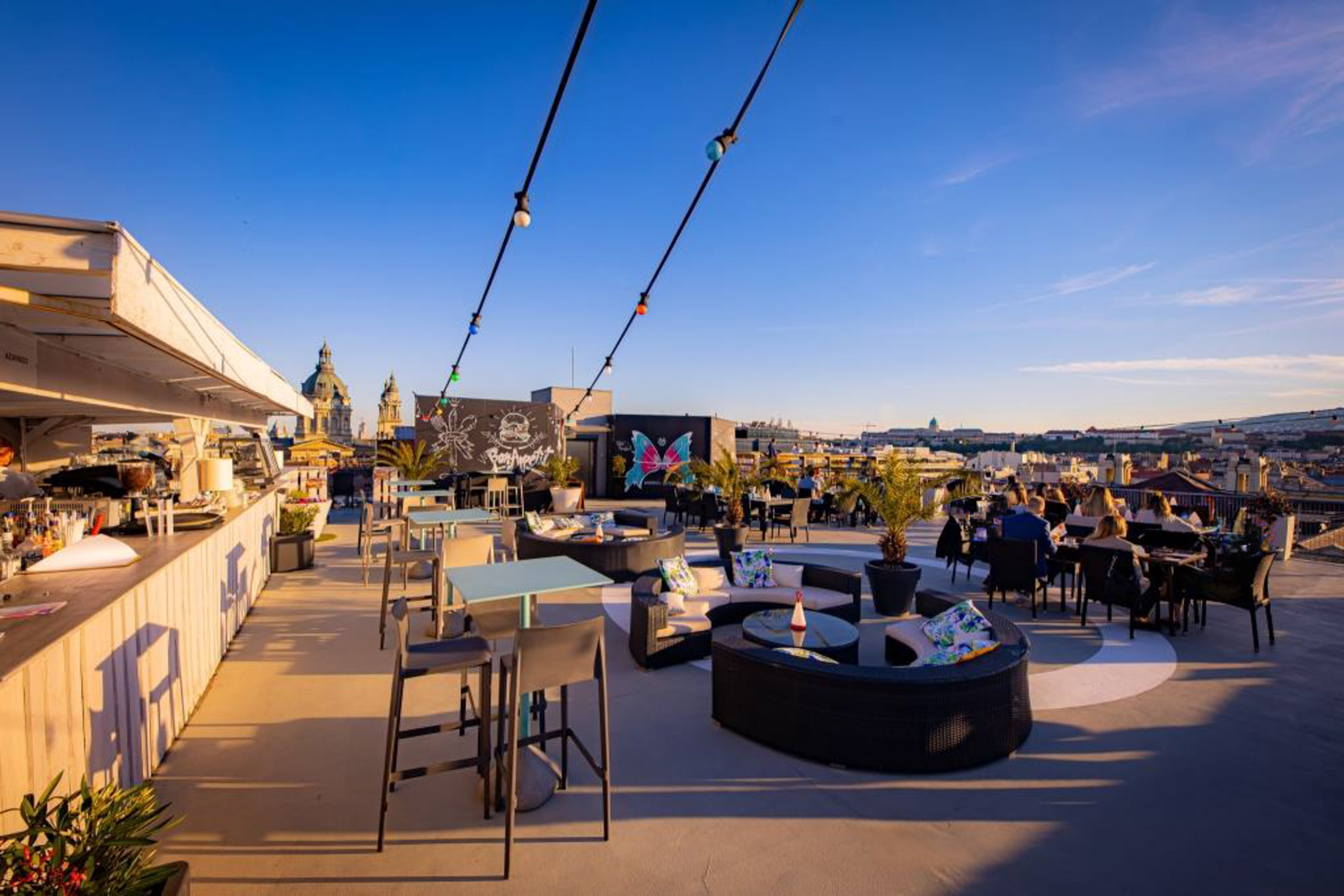 Rooftop bar with outdoor dining, lounge areas, city skyline, and sunset ambiance.