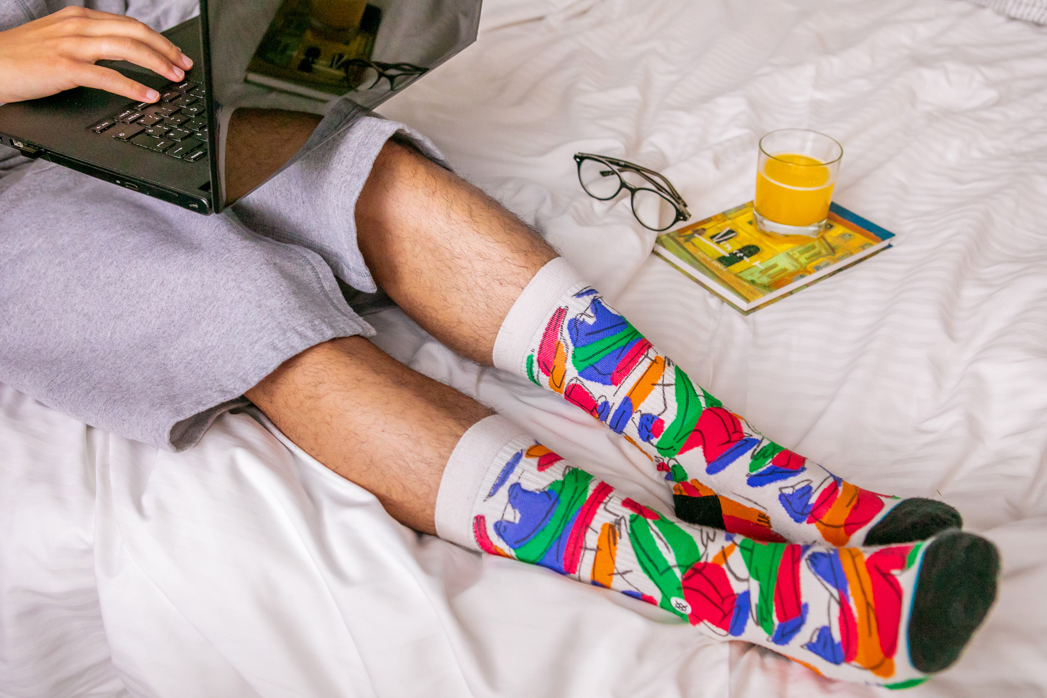 Cozy remote work: person in vibrant socks using laptop on bed with juice.