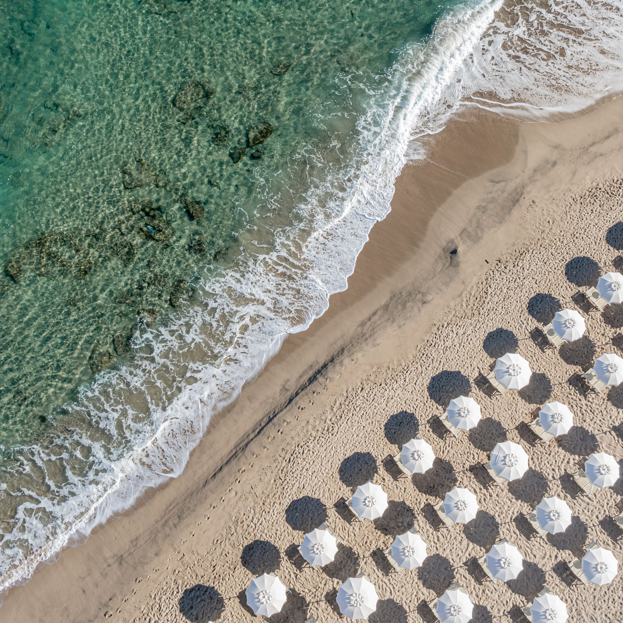 Aerial beach view: turquoise ocean, waves, sandy shore, white umbrellas, sunbeds.