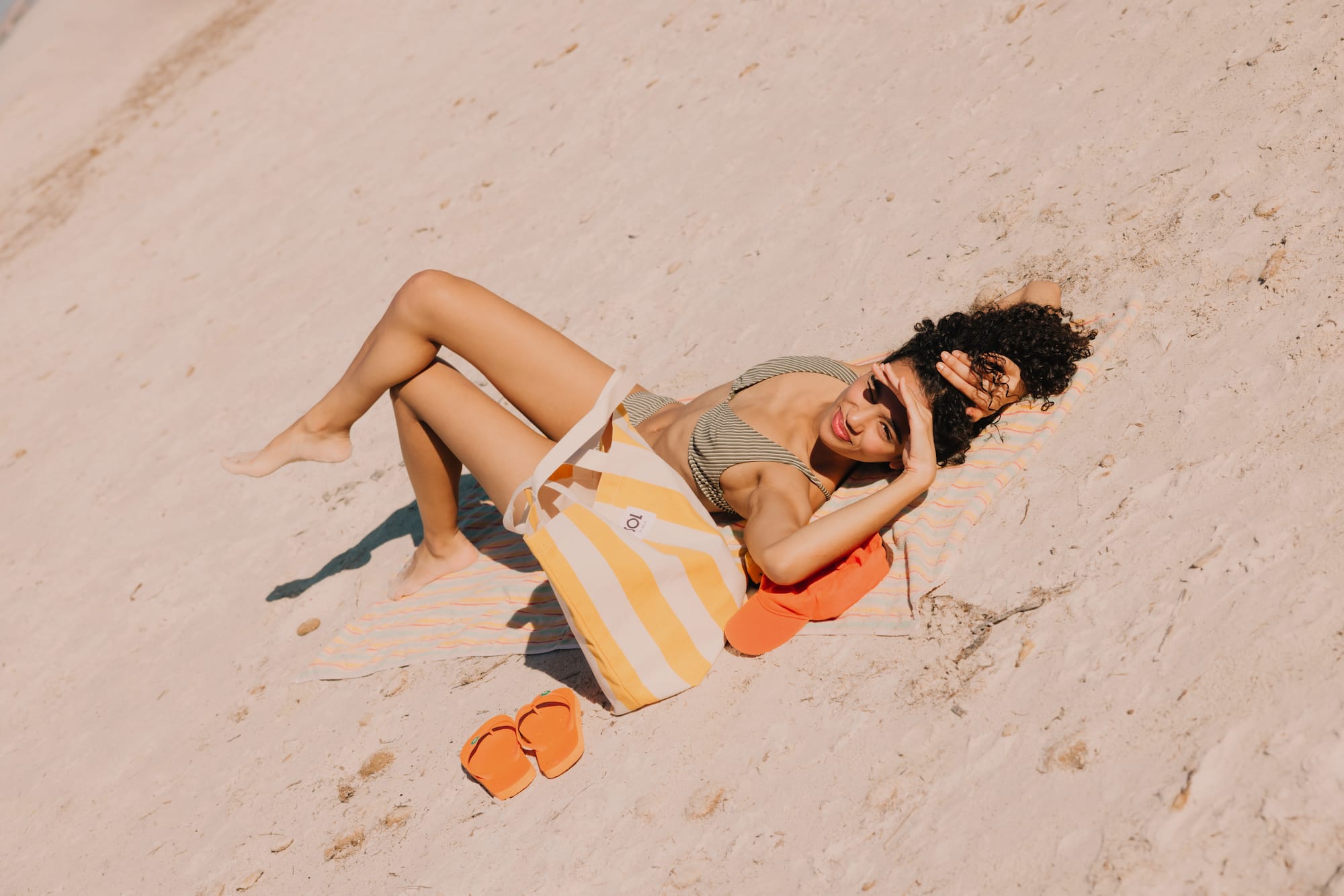 Mujer en bikini a rayas descansando en la playa con bolso y chanclas.
