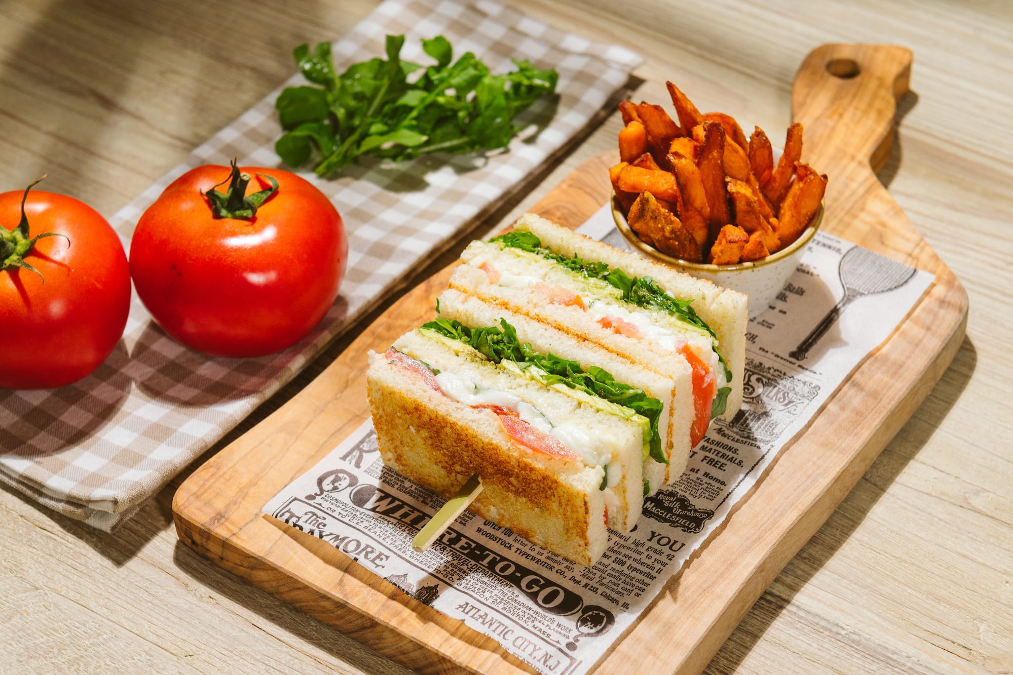 Club sandwich, sweet potato fries, and fresh tomatoes on a wooden board.