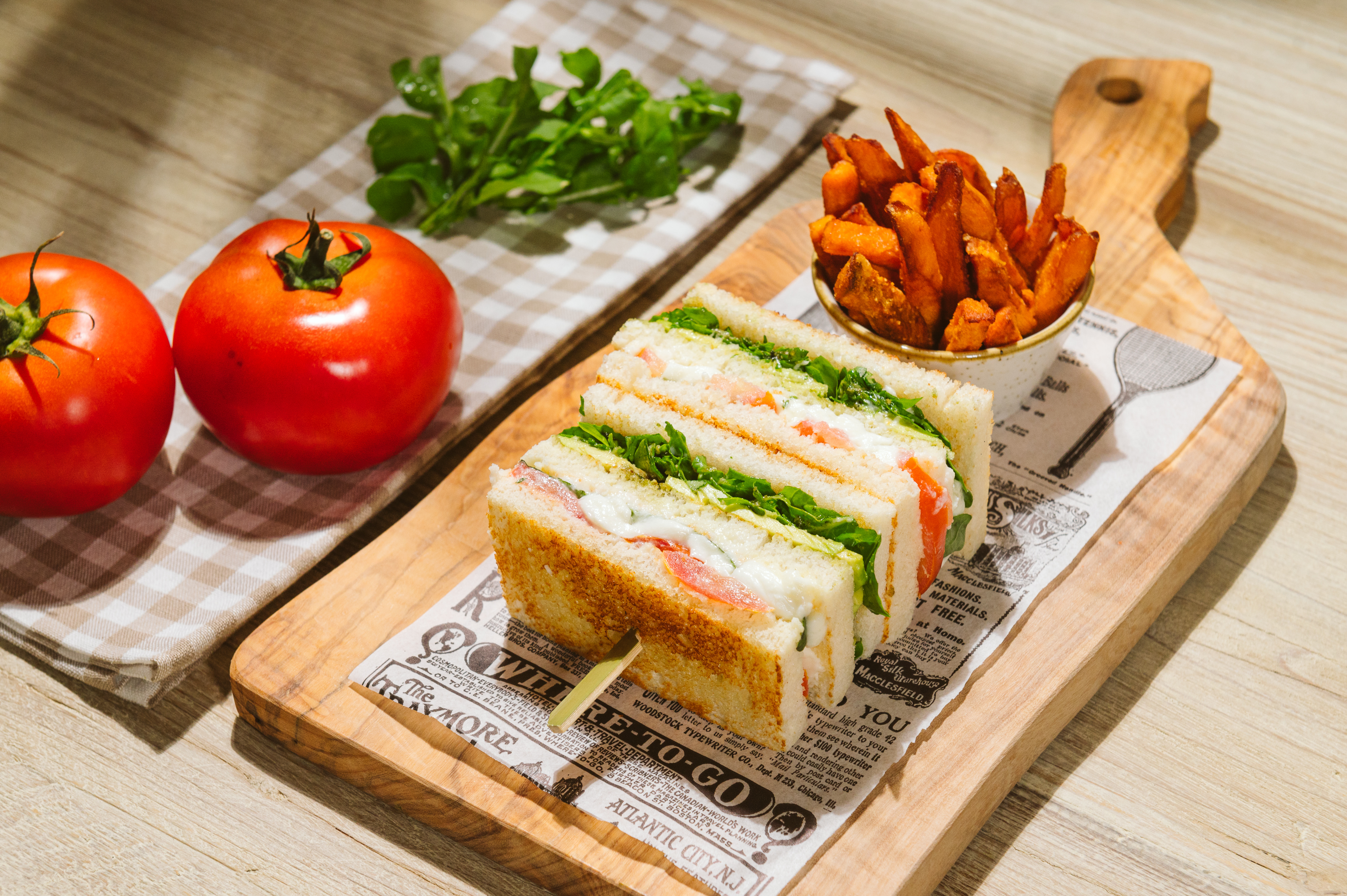 Club sandwich, sweet potato fries, and fresh tomatoes on a wooden board.