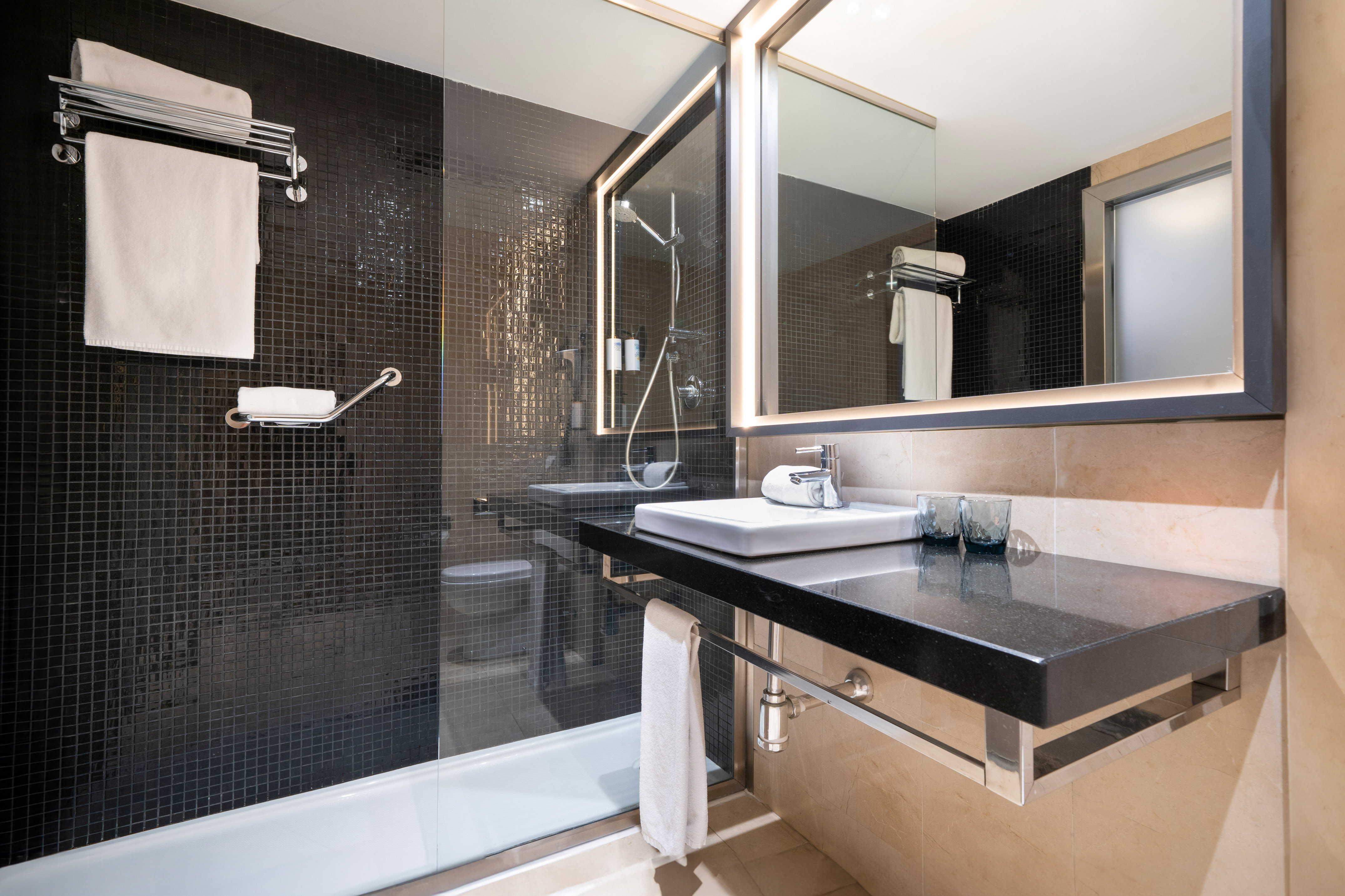 Modern hotel bathroom with black tiled shower, white bathtub, and vanity.