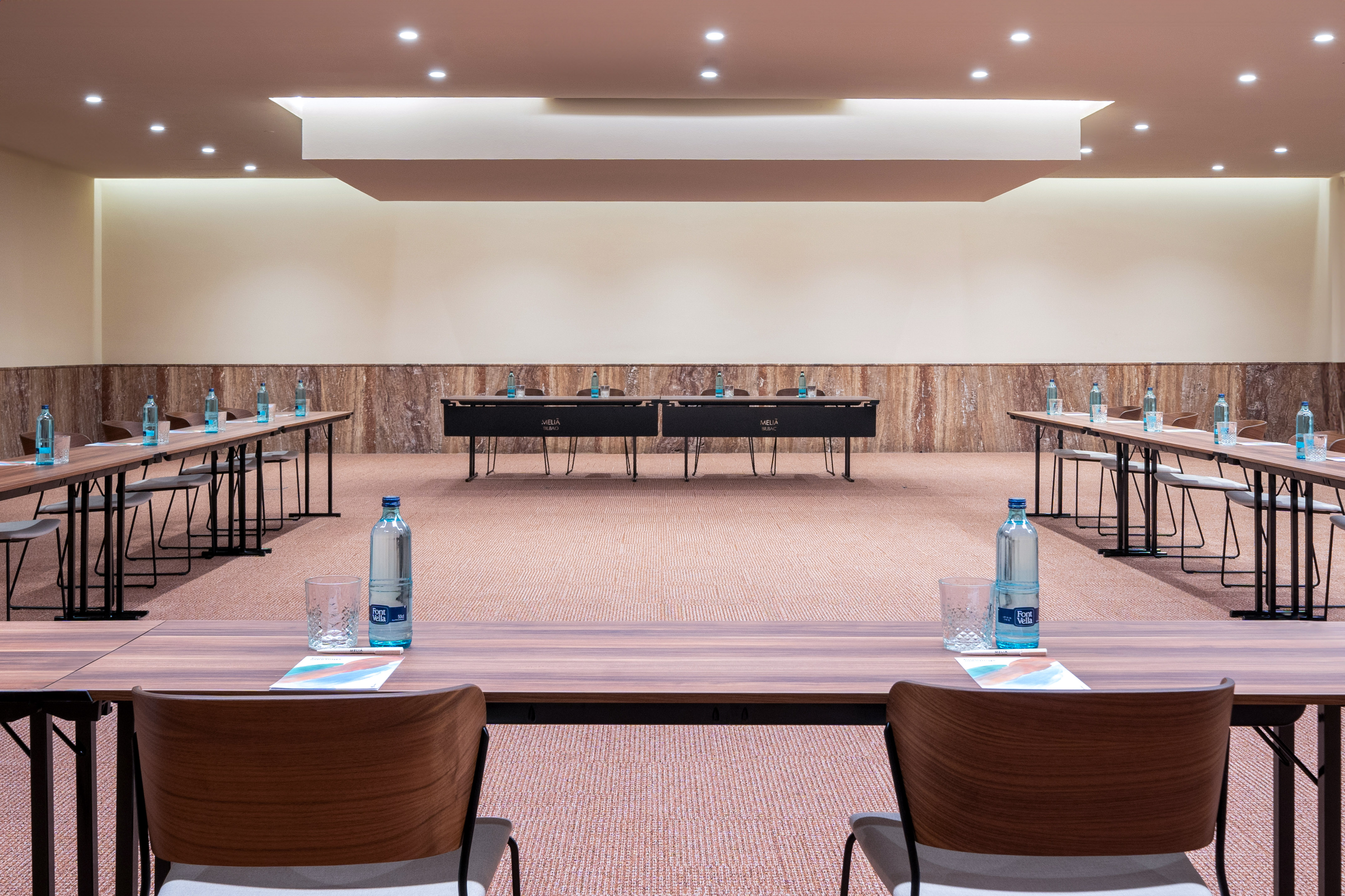 Empty hotel conference room with multiple tables, chairs, and bottled water setup.