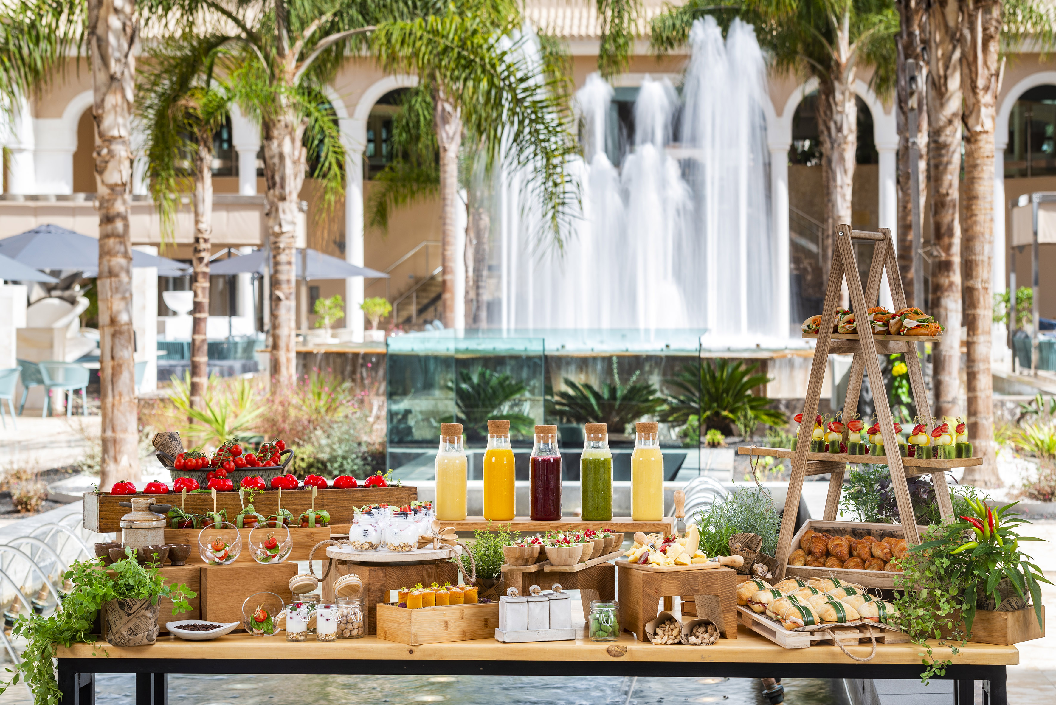 Outdoor resort buffet with fresh juices, snacks, and fruit by a grand fountain.