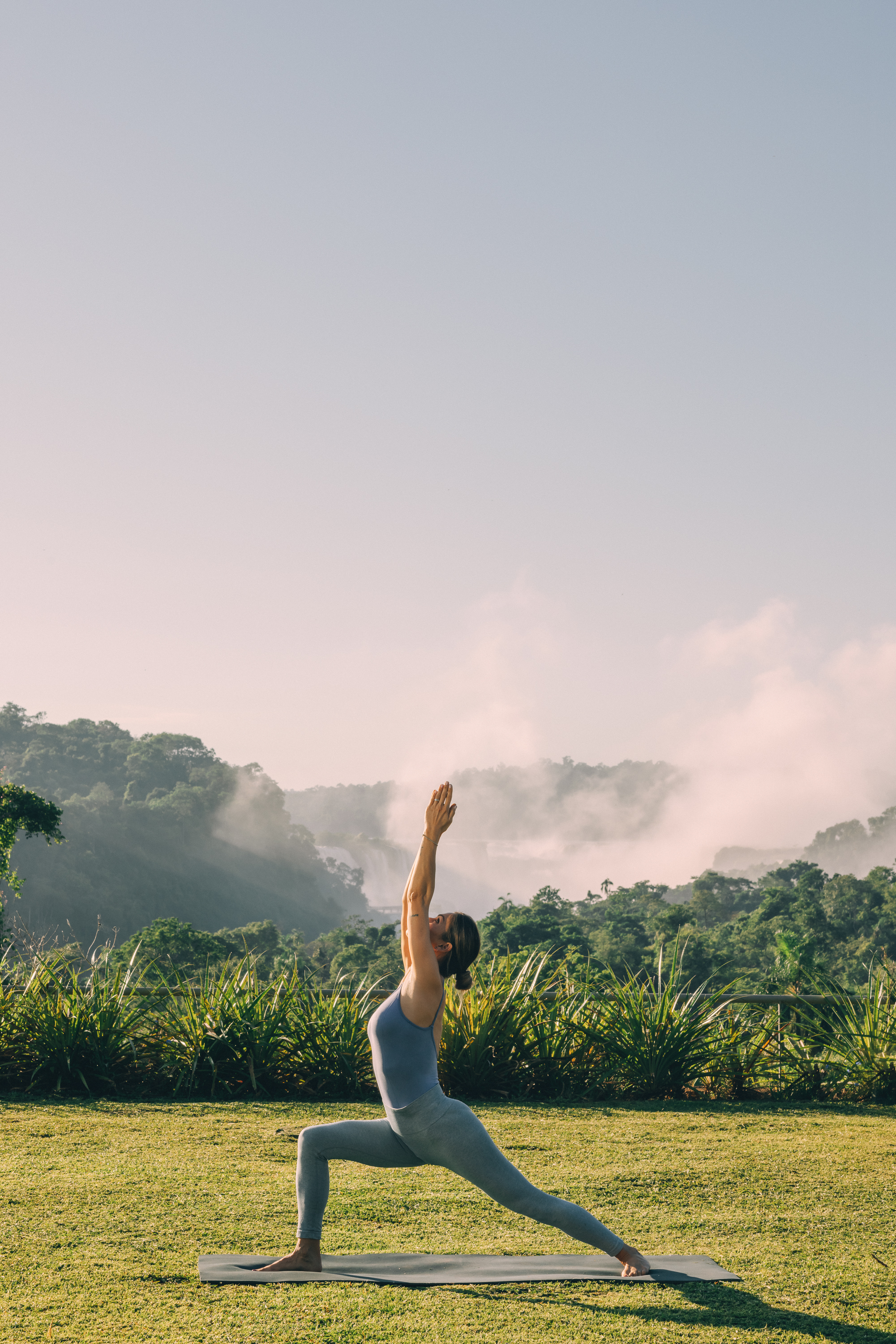 Woman in yoga warrior pose on mat, misty waterfall and lush greenery.