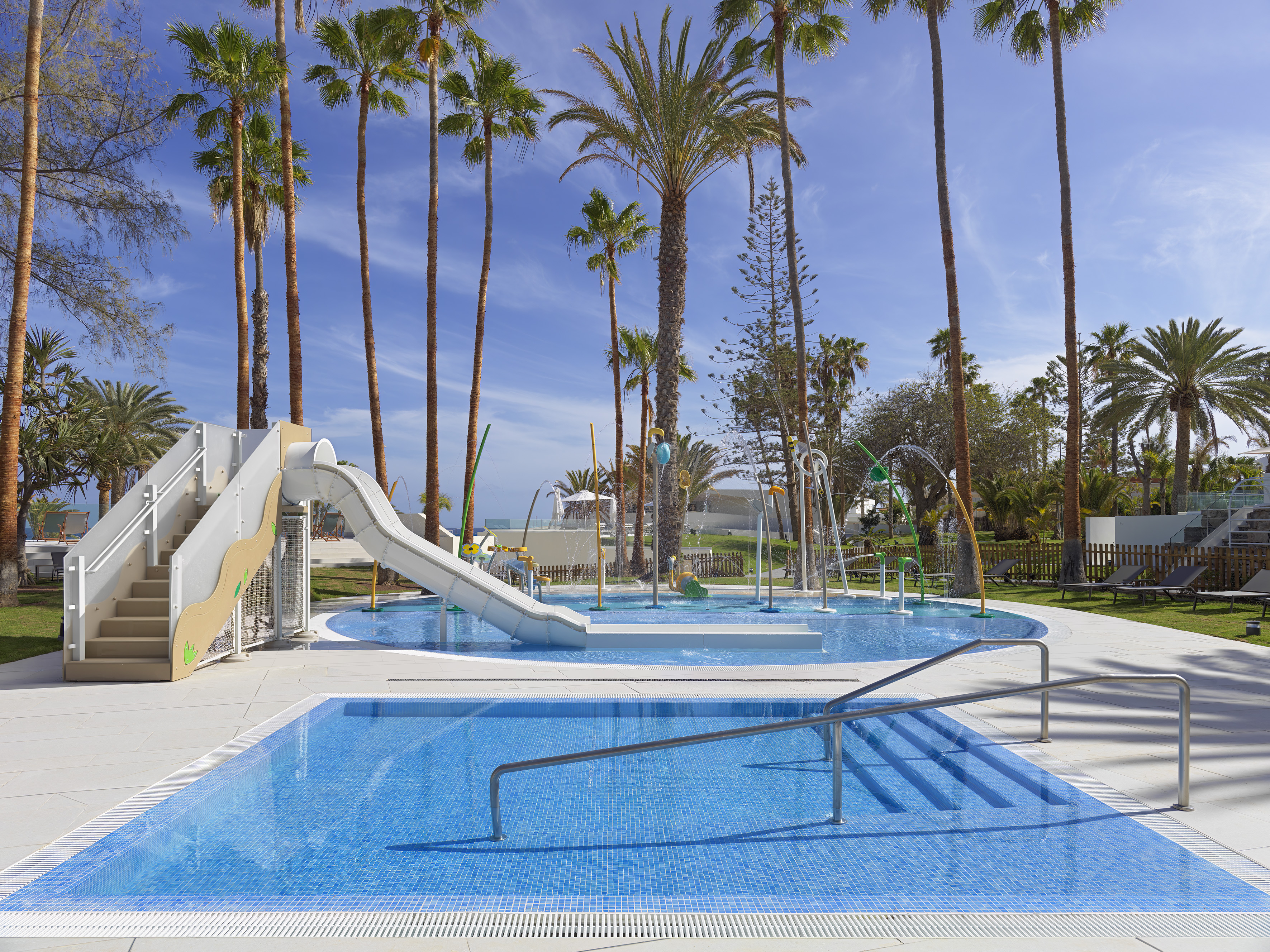 Kids' splash park with water slide, shallow pool, and palm trees under a blue sky.