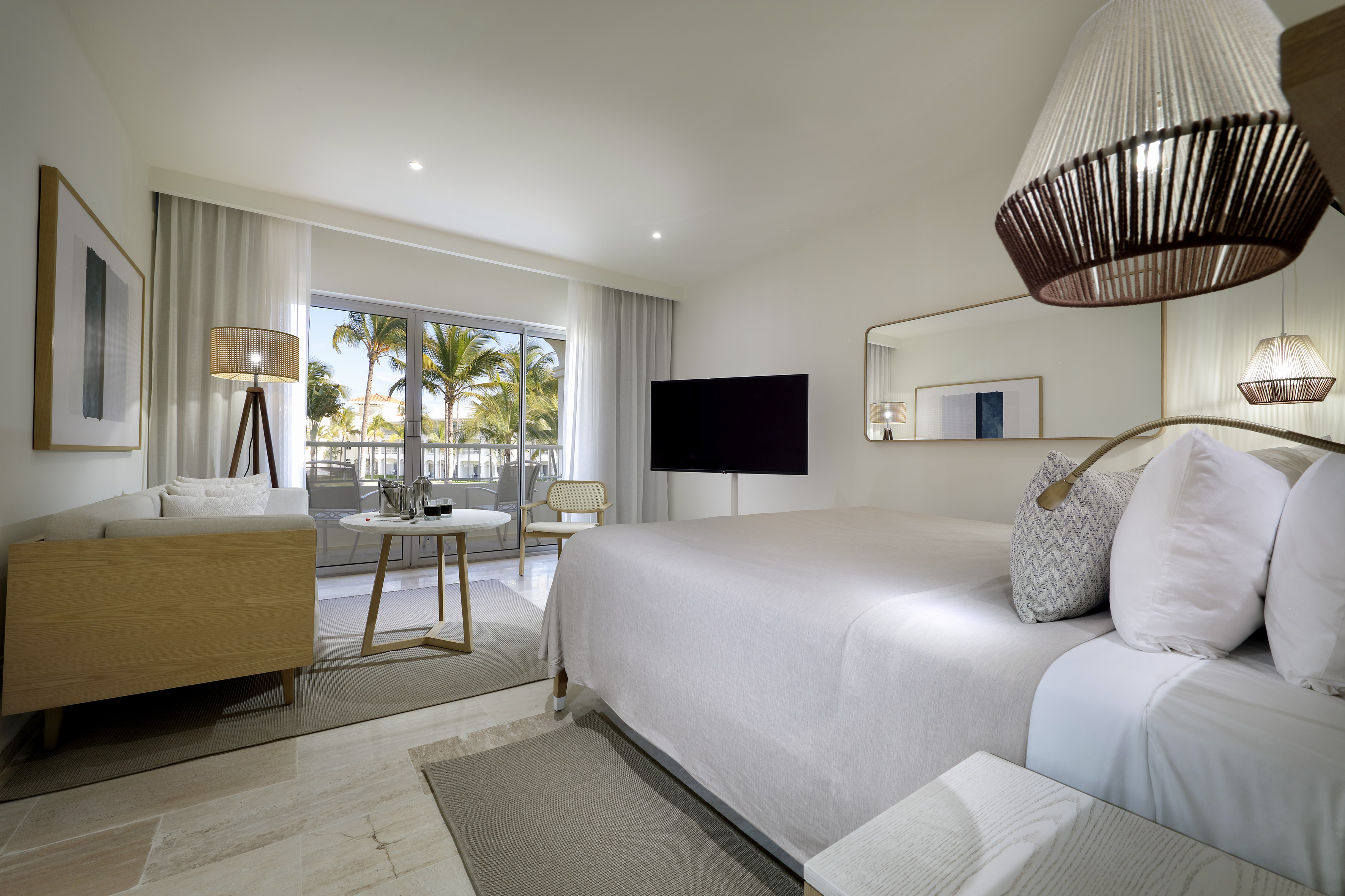 Modern resort room with king bed, TV, sofa, and tropical balcony view.
