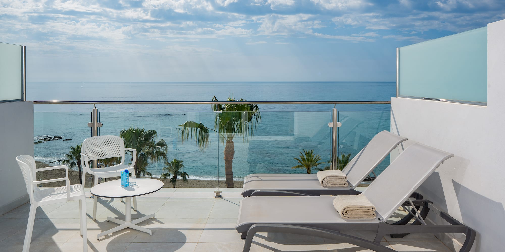 Hotel balcony with ocean view, beach, palm trees, and lounge chairs.