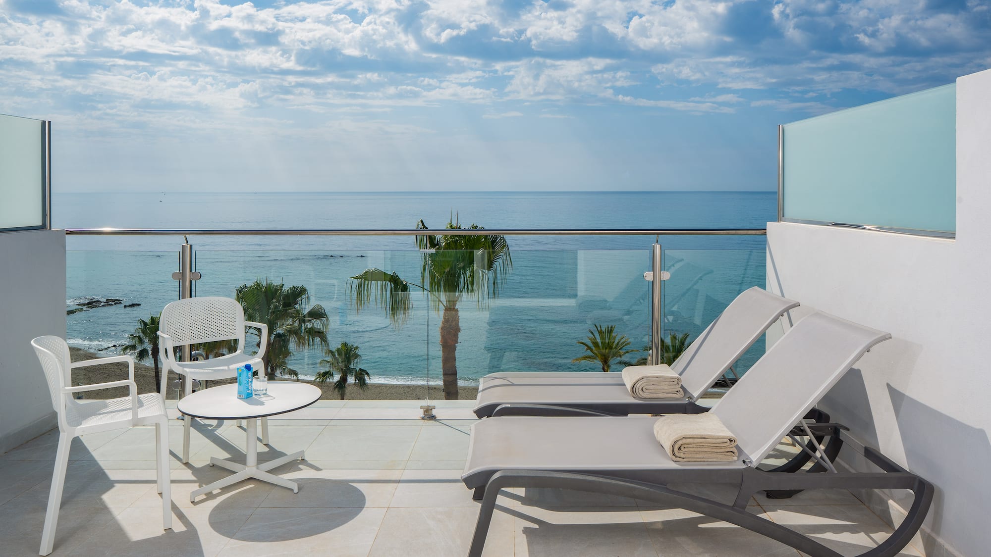 Hotel balcony with ocean view, sun loungers, palm trees, and beach.