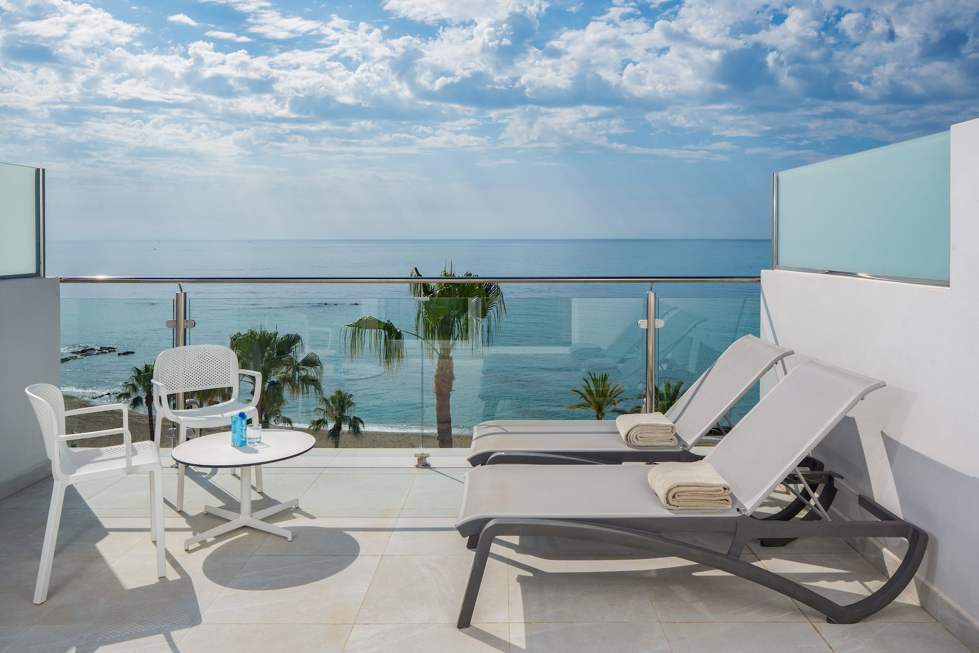 Hotel balcony with ocean view, beach, palm trees, and lounge chairs.