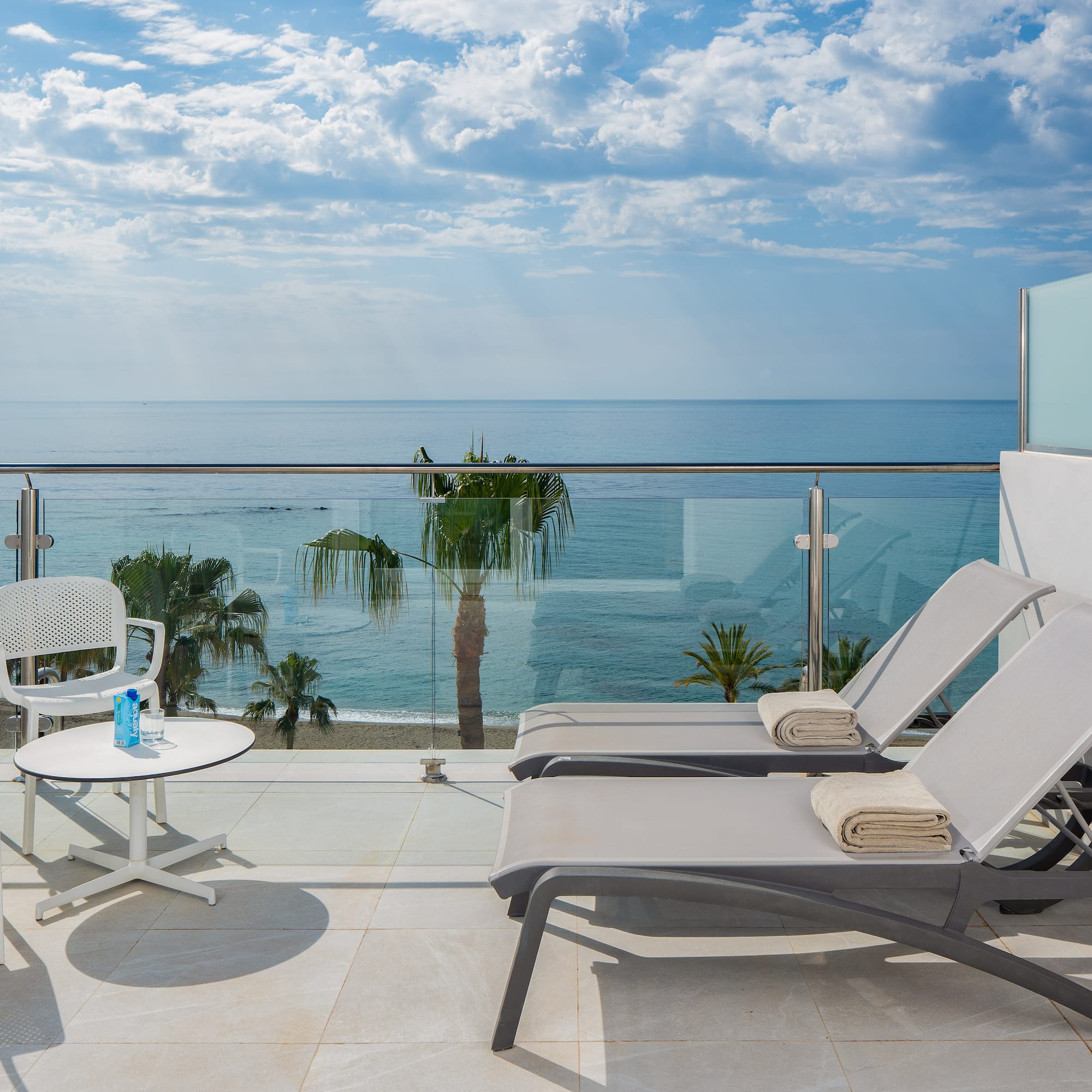 Hotel balcony with ocean view, beach, palm trees, and lounge chairs.