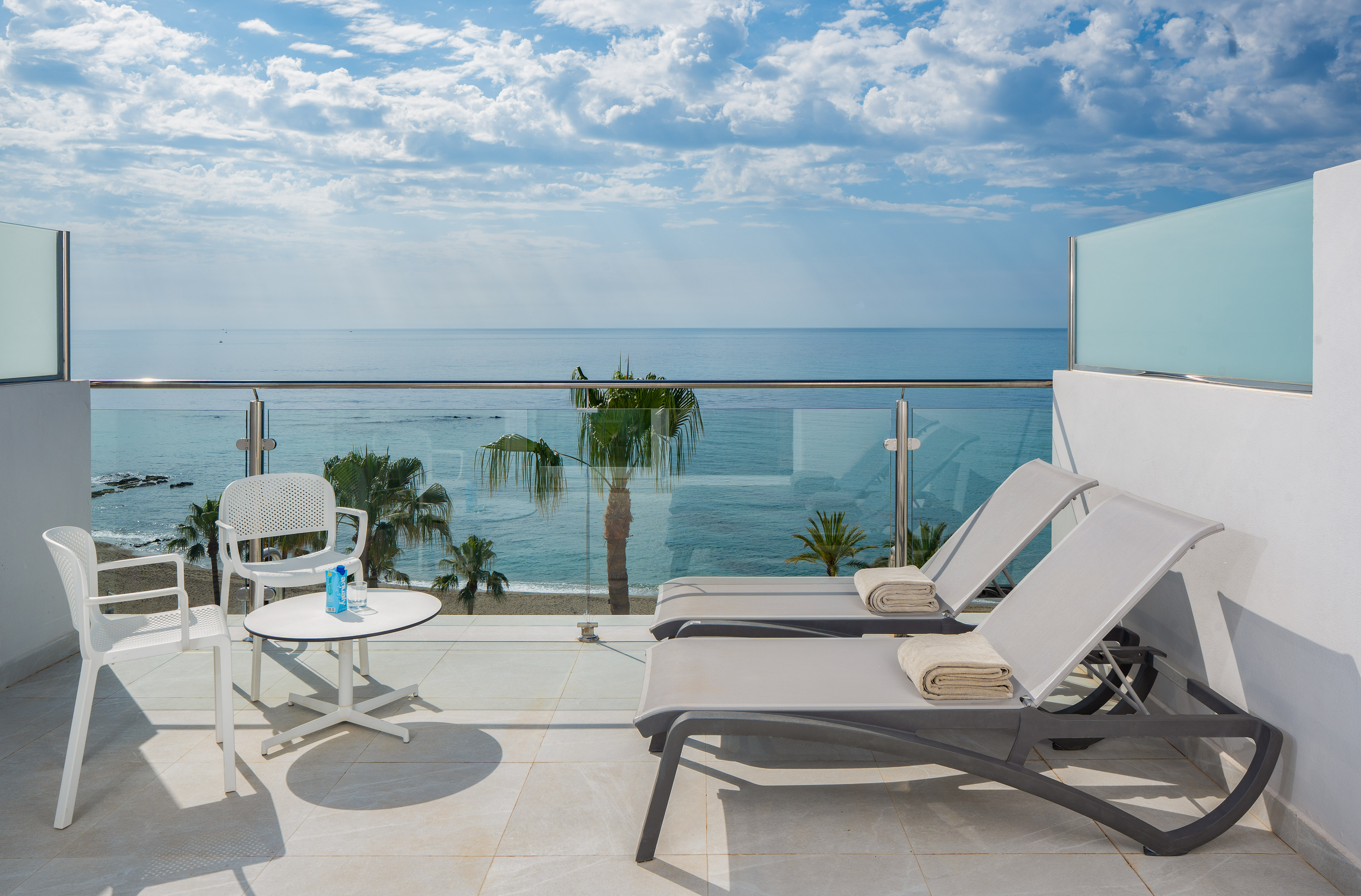 Hotel balcony with ocean view, sun loungers, palm trees, and beach.