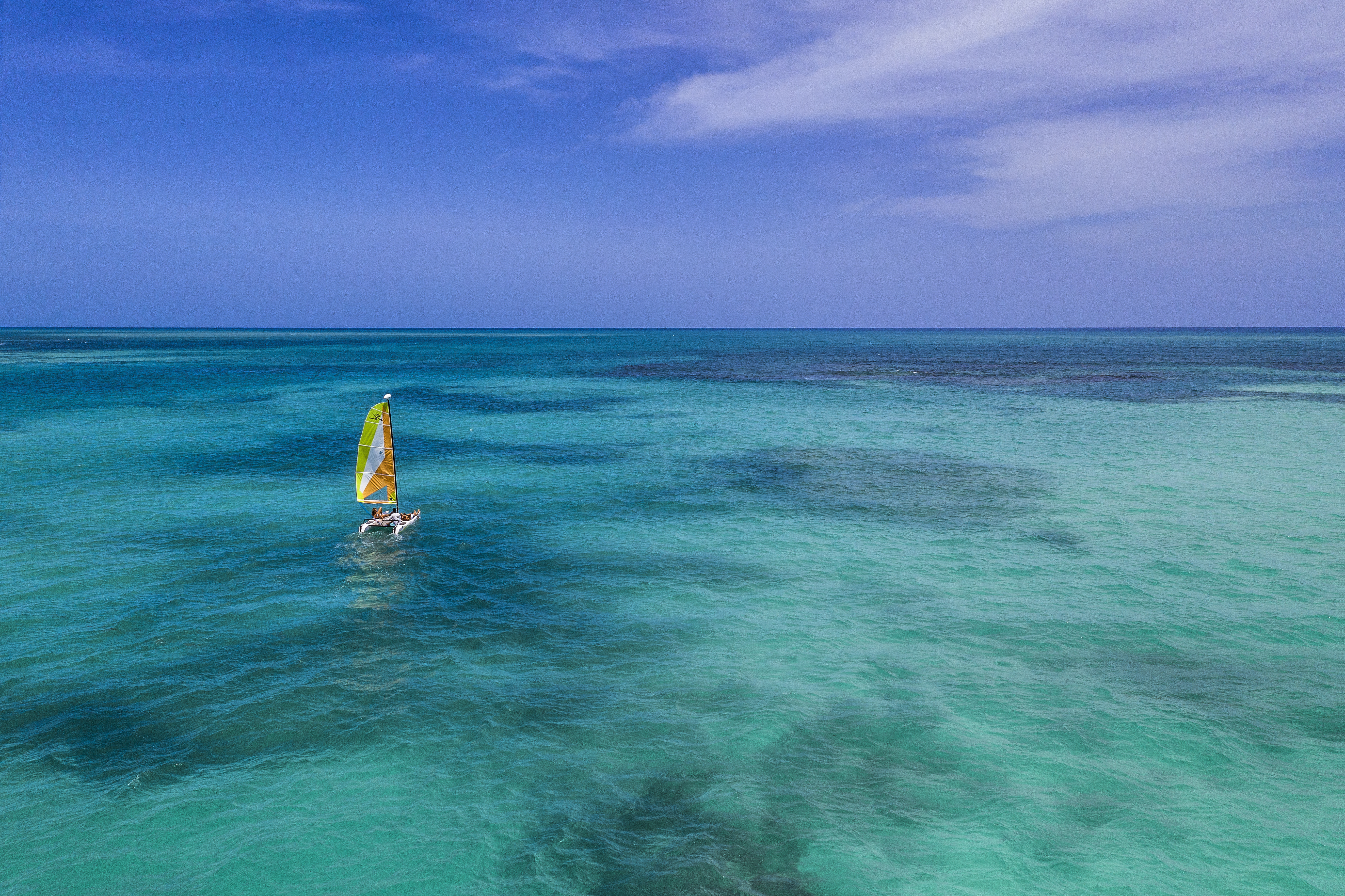 Catamaran with yellow sail glides on clear blue tropical ocean.