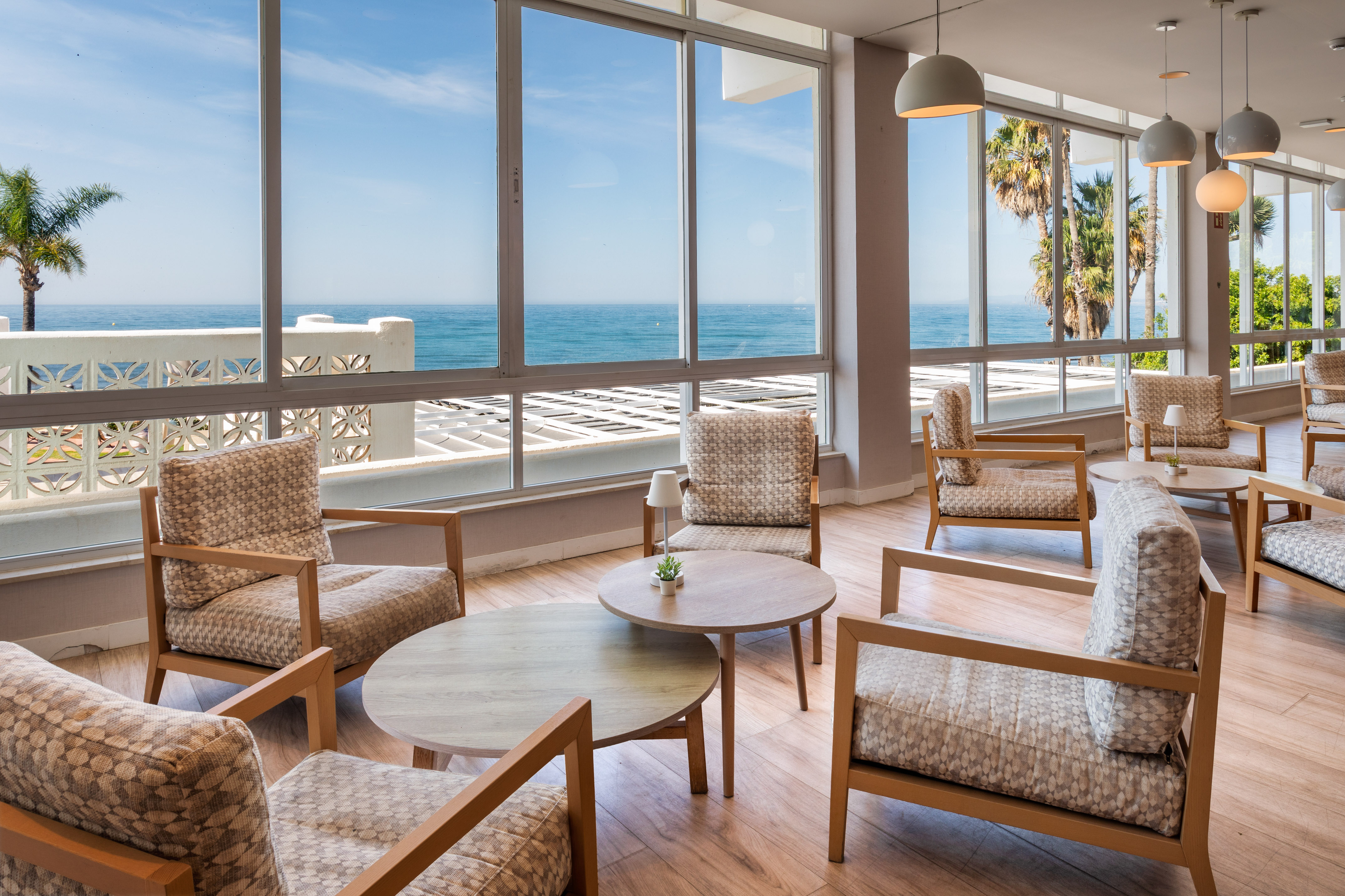 Hotel lounge with panoramic ocean view, modern seating, and palm trees.