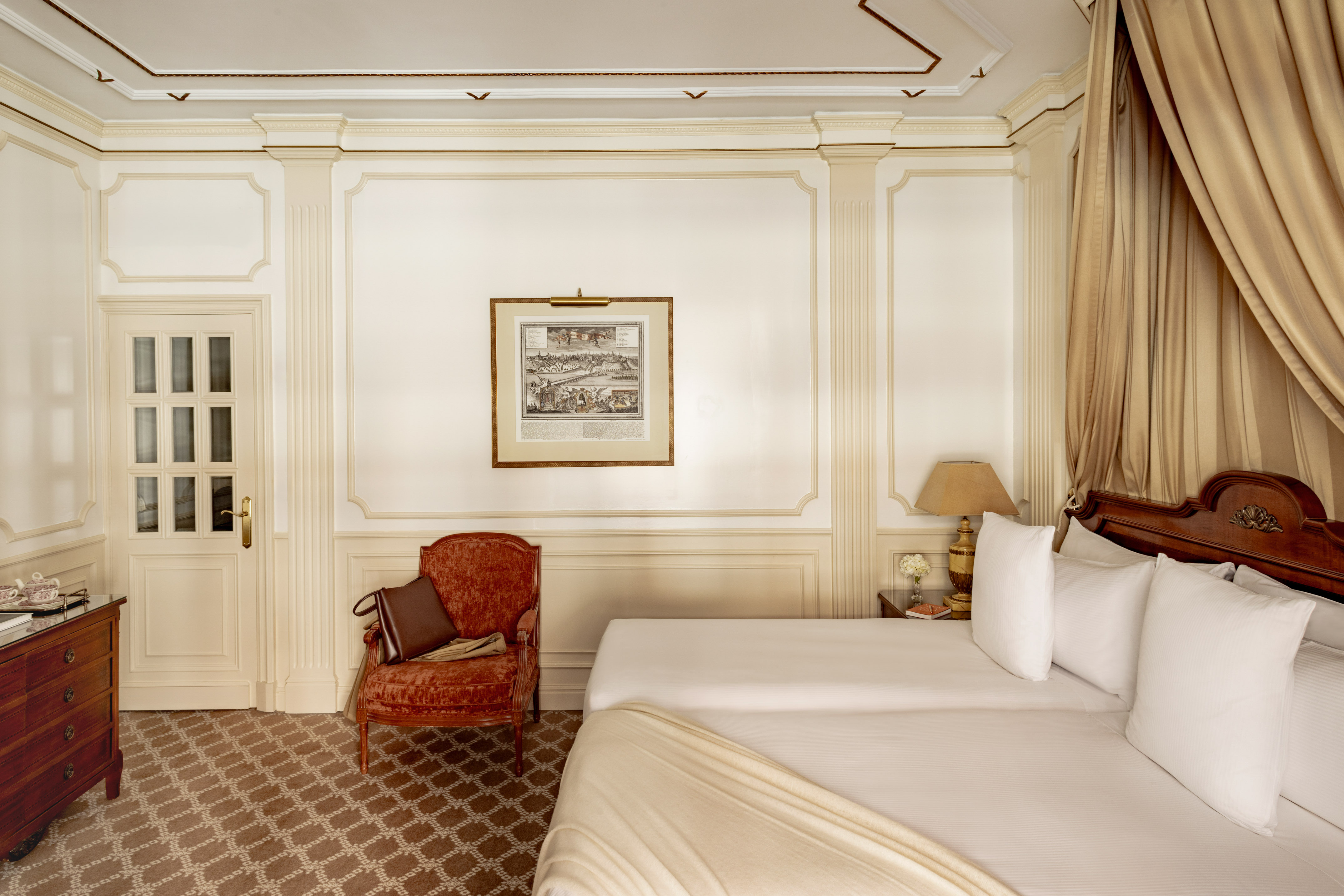 Elegant classic hotel bedroom with ornate molding, twin beds, and velvet armchair.