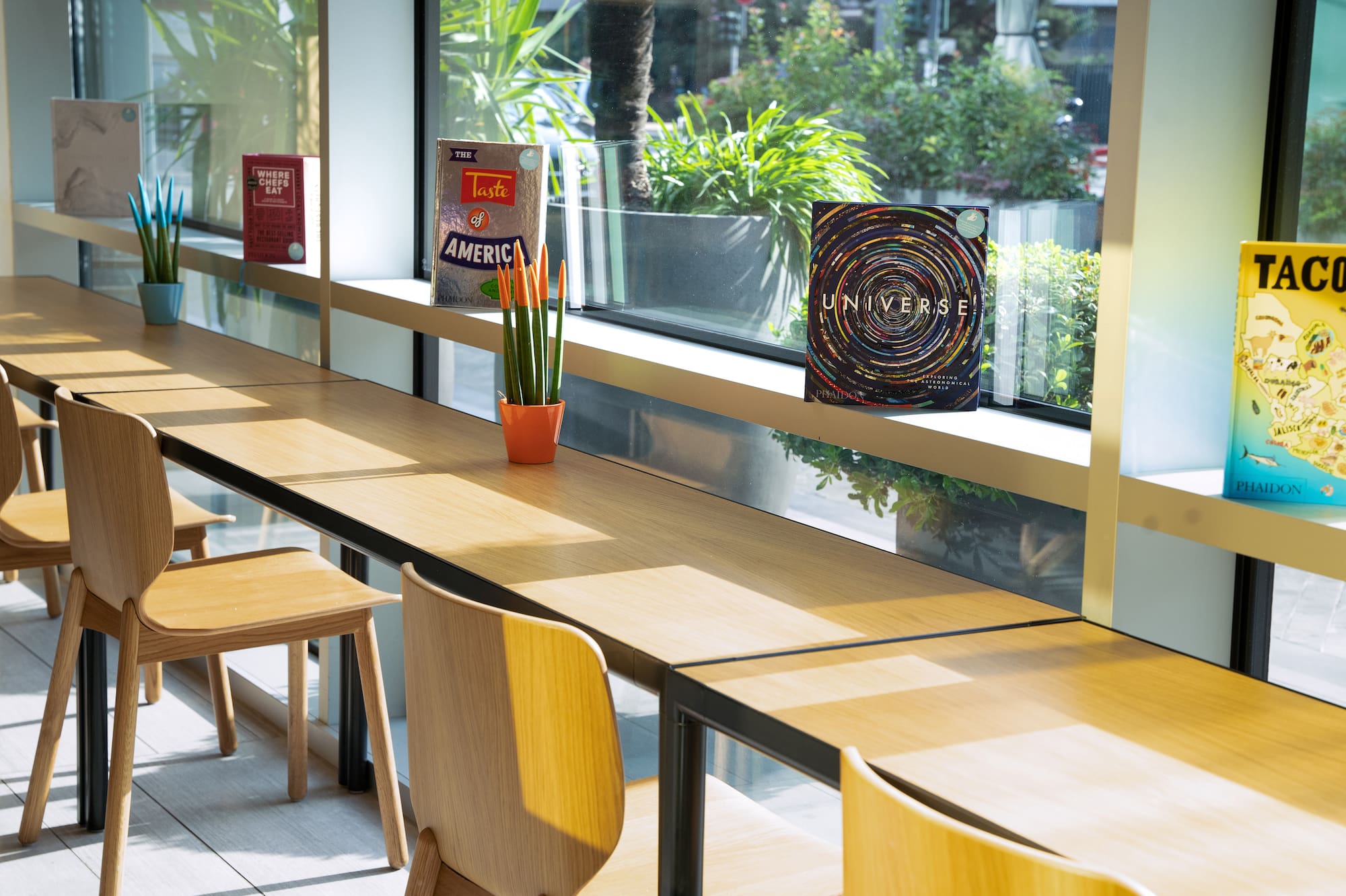 Modern cafe seating, wooden tables, Phaidon books, plants by bright window.