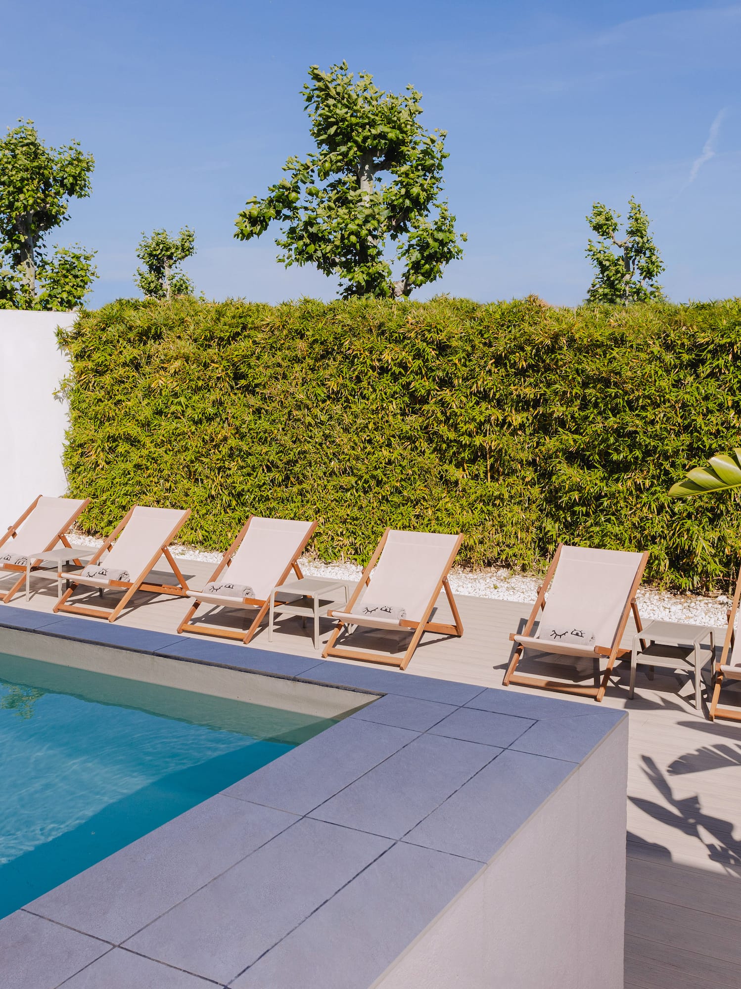 a pool with lounge chairs and a wall with a hedge and a blue sky