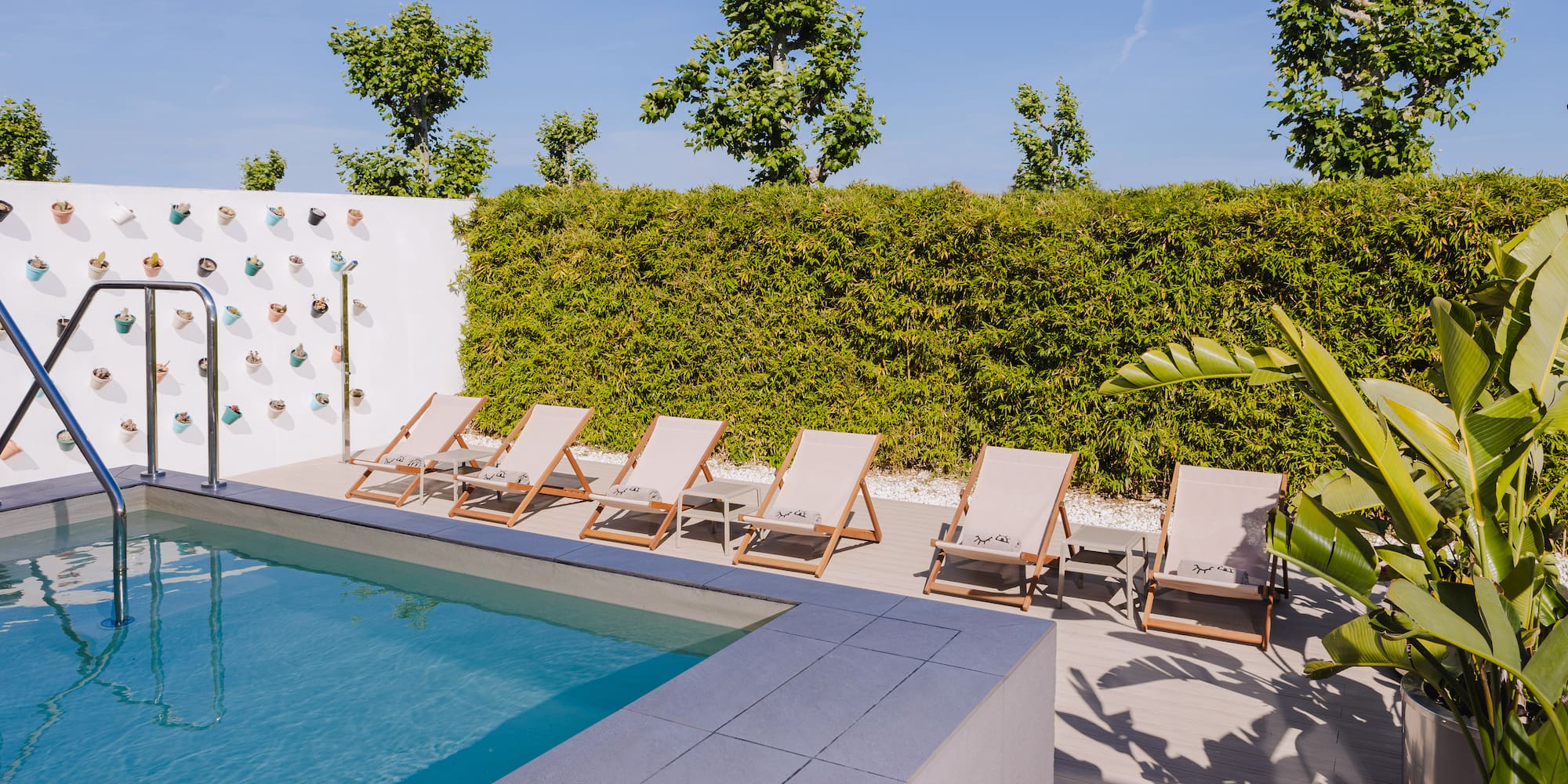a pool with lounge chairs and a wall with a hedge and a blue sky