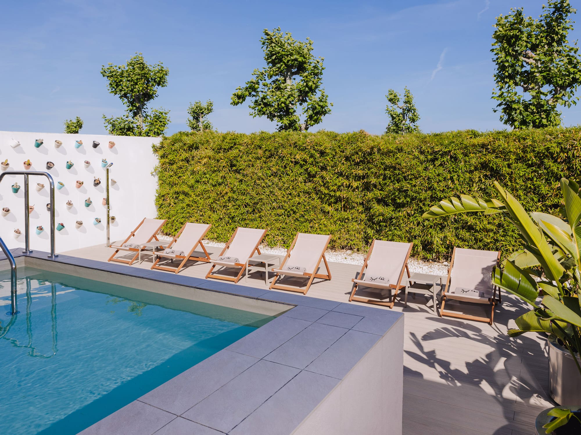 a pool with lounge chairs and a wall with a hedge and a blue sky