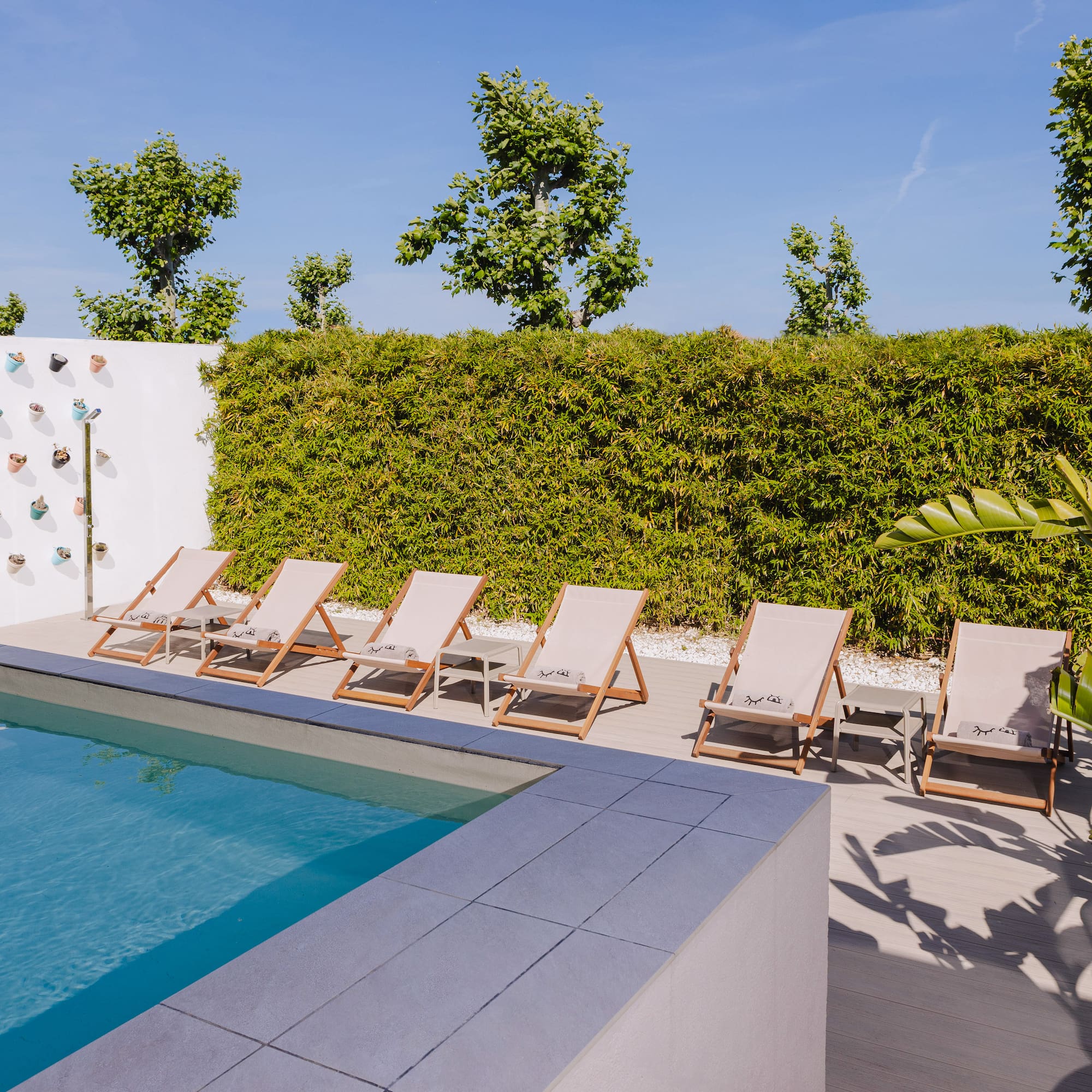 a pool with lounge chairs and a wall with a hedge and a blue sky