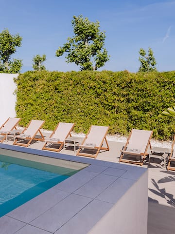 a pool with lounge chairs and a wall with a hedge and a blue sky