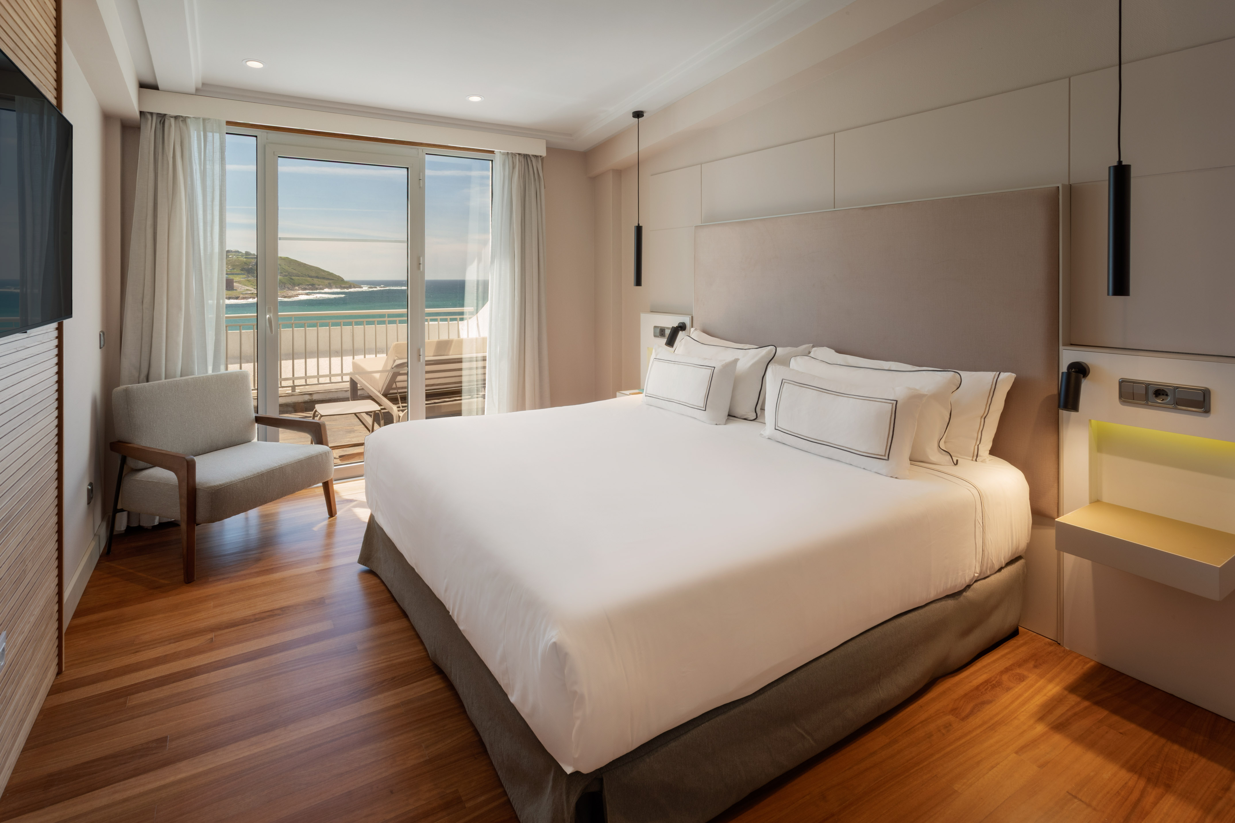 Modern hotel bedroom with large bed, hardwood floor, and ocean view balcony.