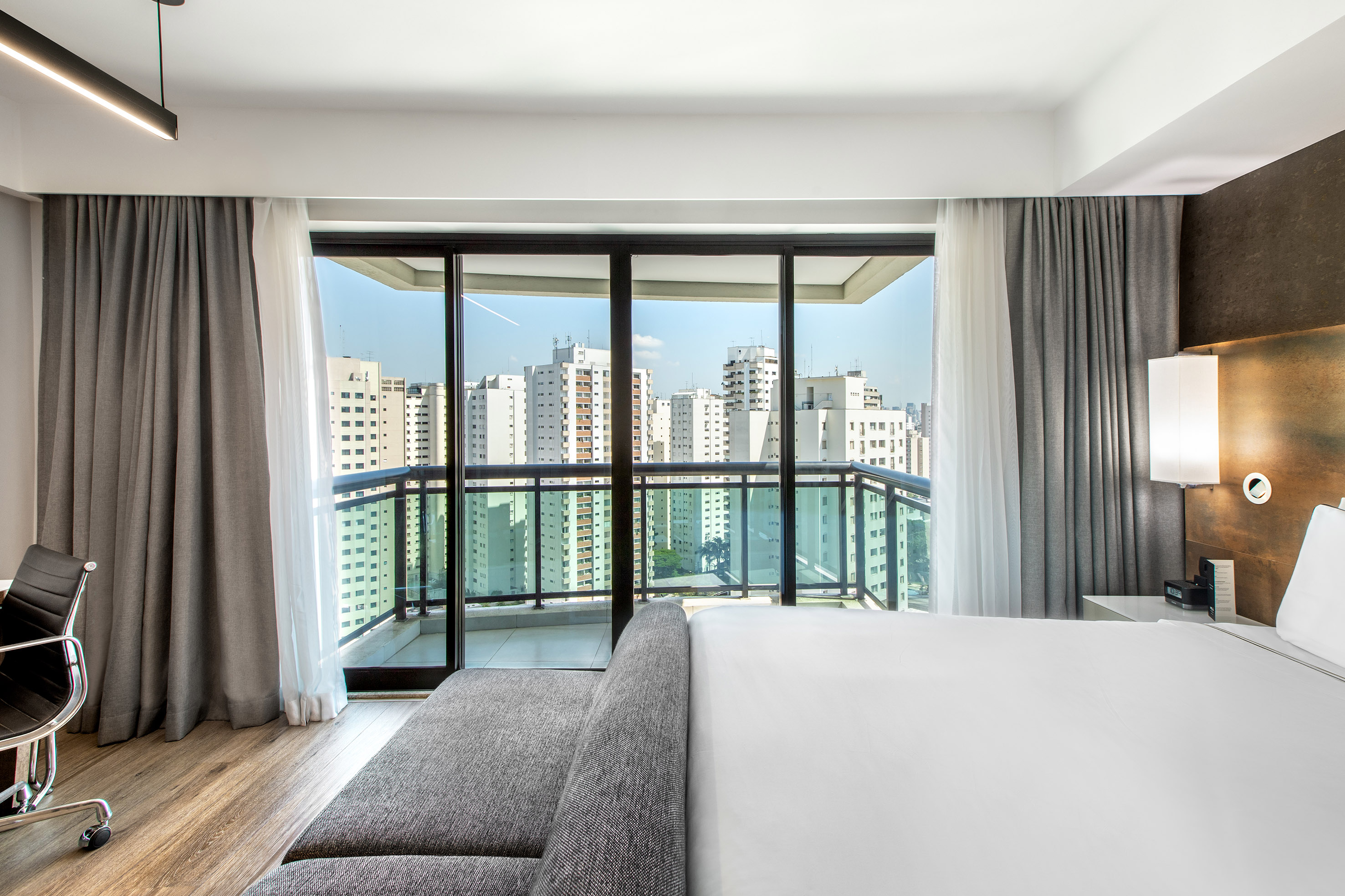 Modern hotel room with balcony, city skyline view, bed, and grey sofa.