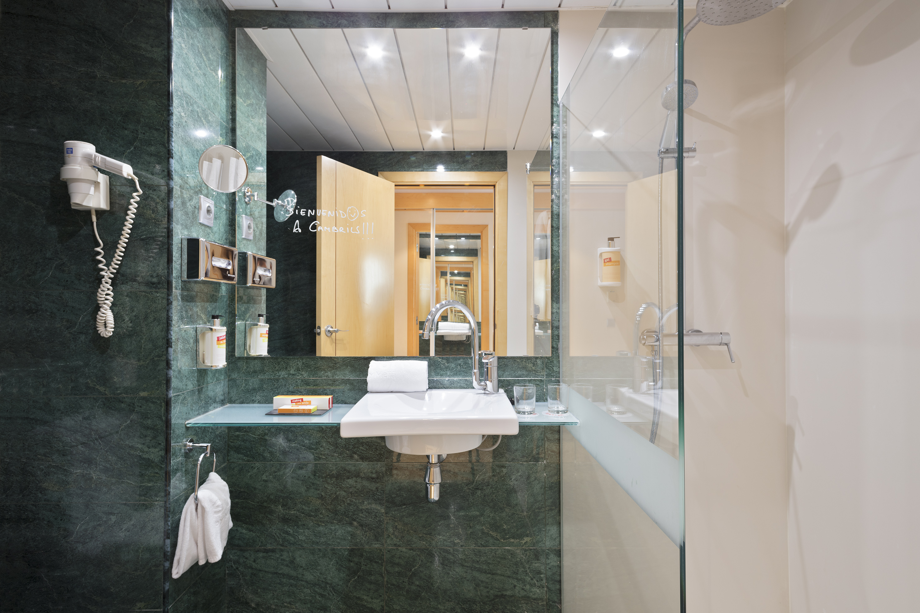 Modern hotel bathroom with green marble, white sink, mirror, and glass shower.