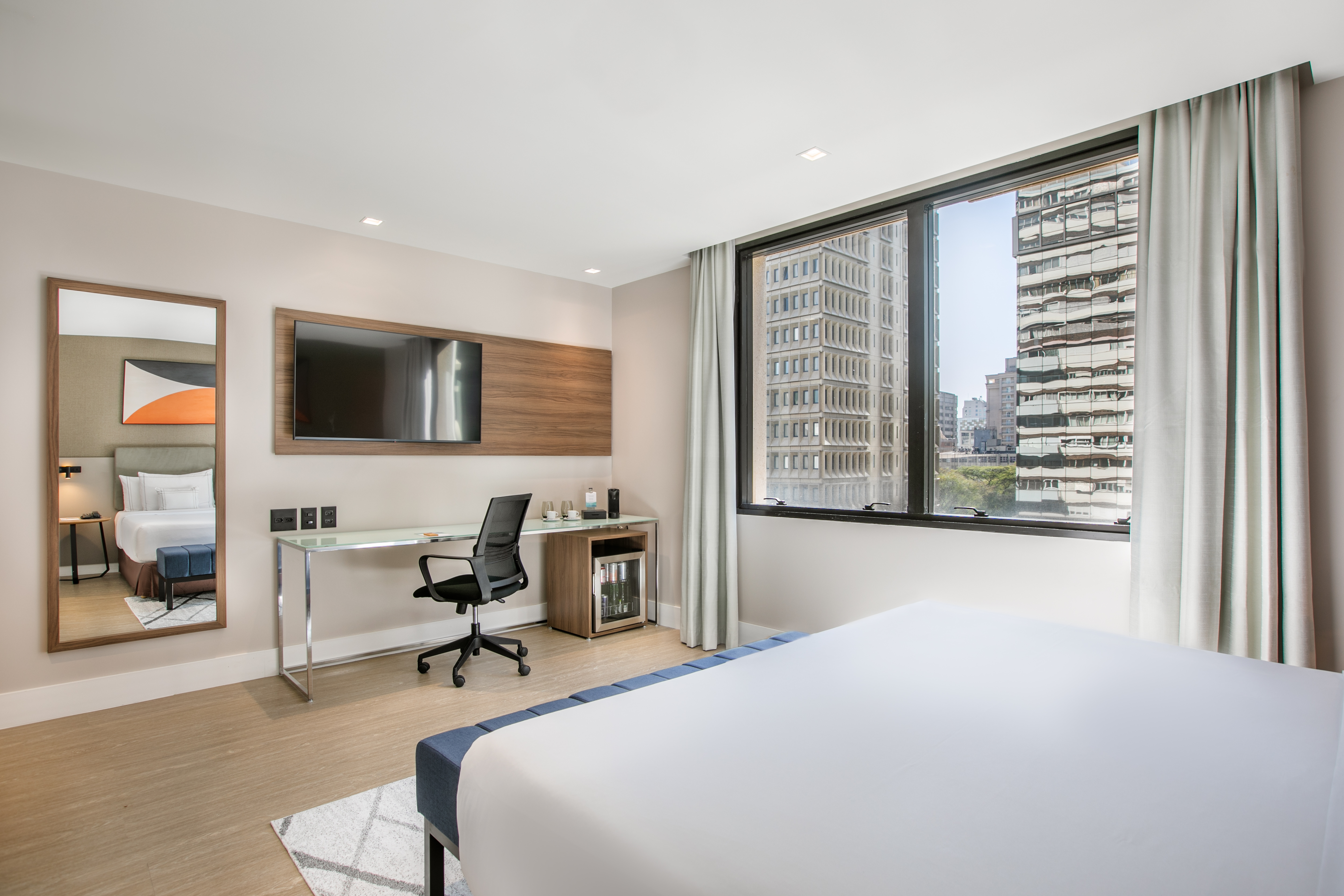 Modern hotel room with bed, desk, TV, and city view through large window.