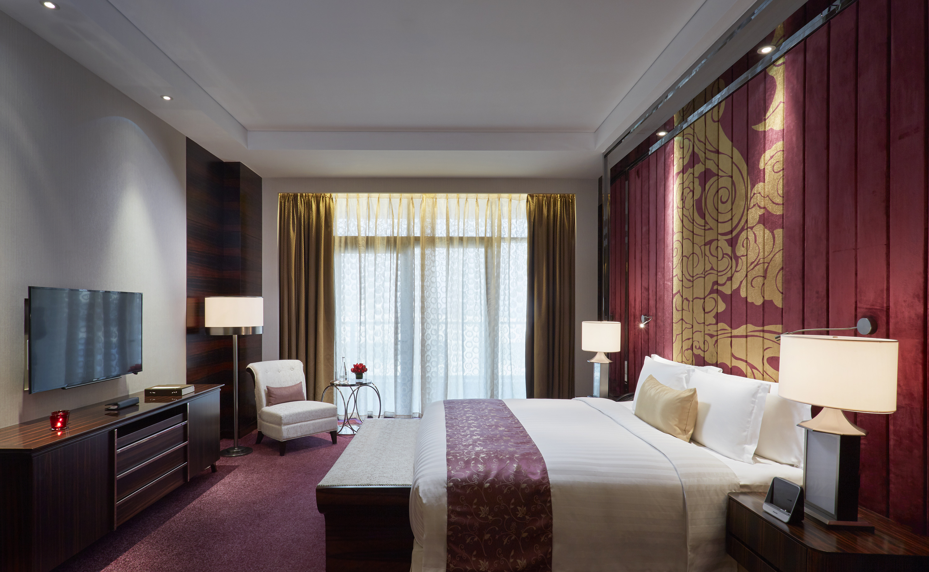 Luxury hotel bedroom with king bed, ornate red gold headboard, large window.