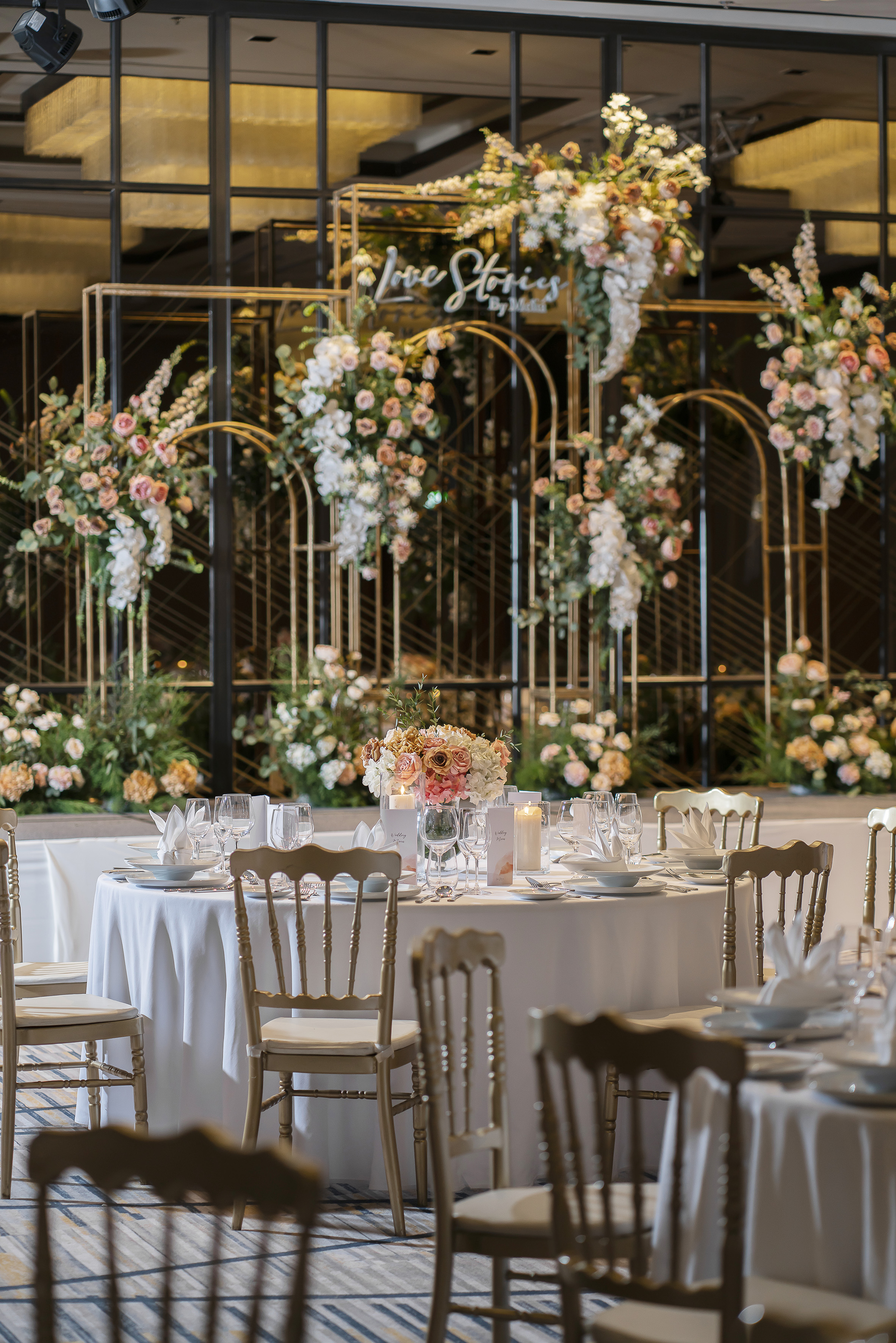 Wedding reception tables with floral centerpieces and 'Love Stories' gold arch.