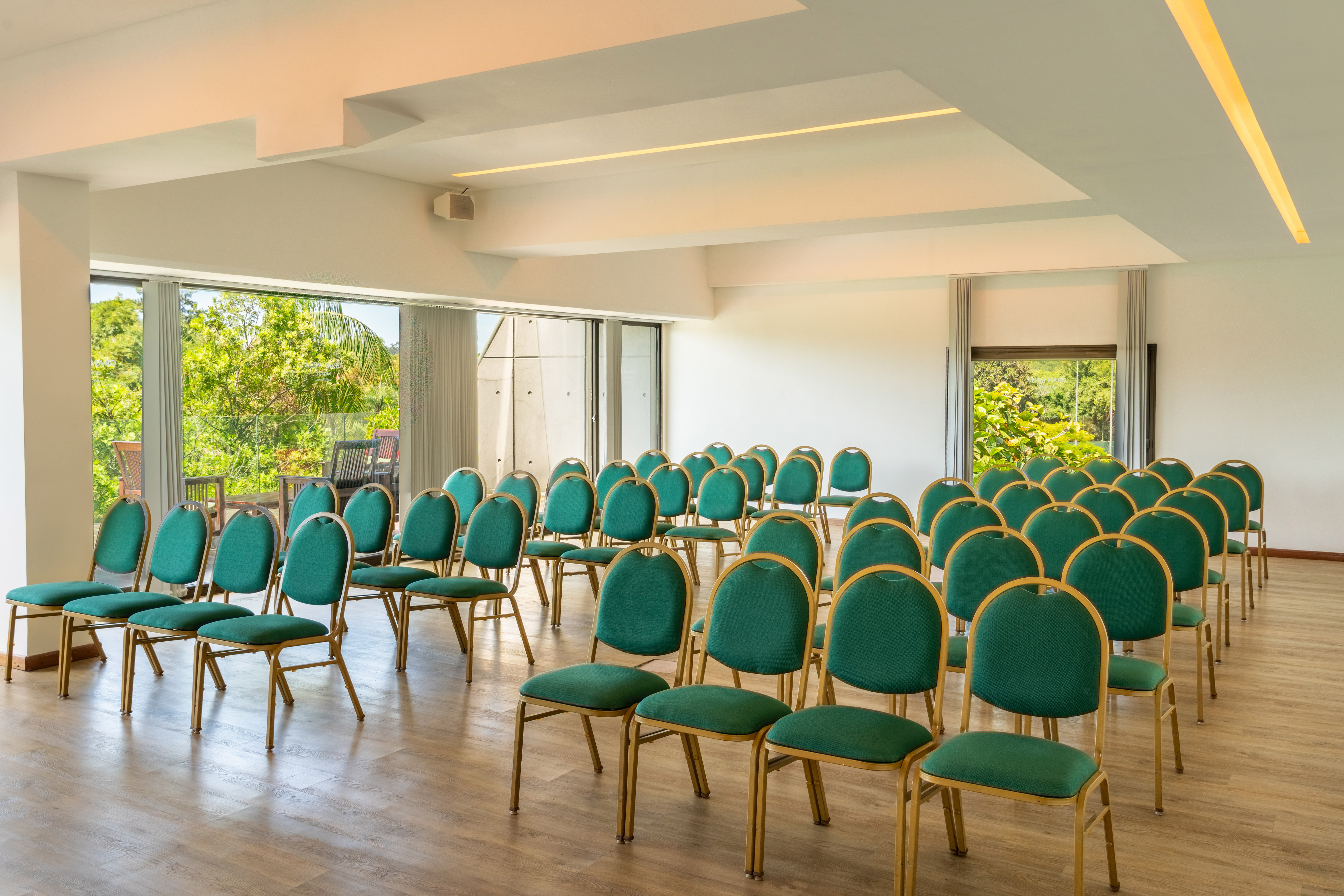 Conference room with rows of green chairs, large windows, and lush outdoor view.