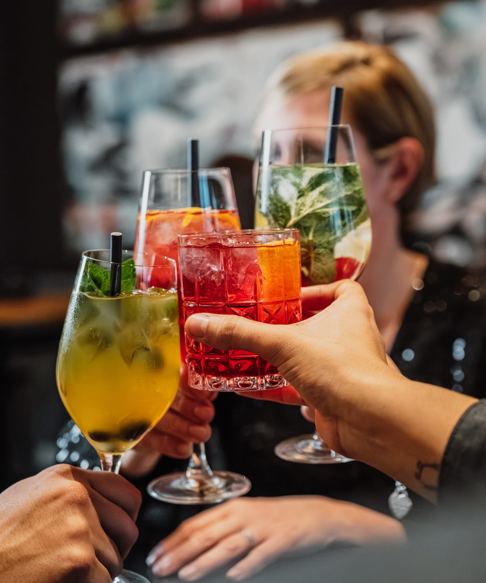 Hands toasting vibrant cocktails with ice, fruit, and mint garnishes.
