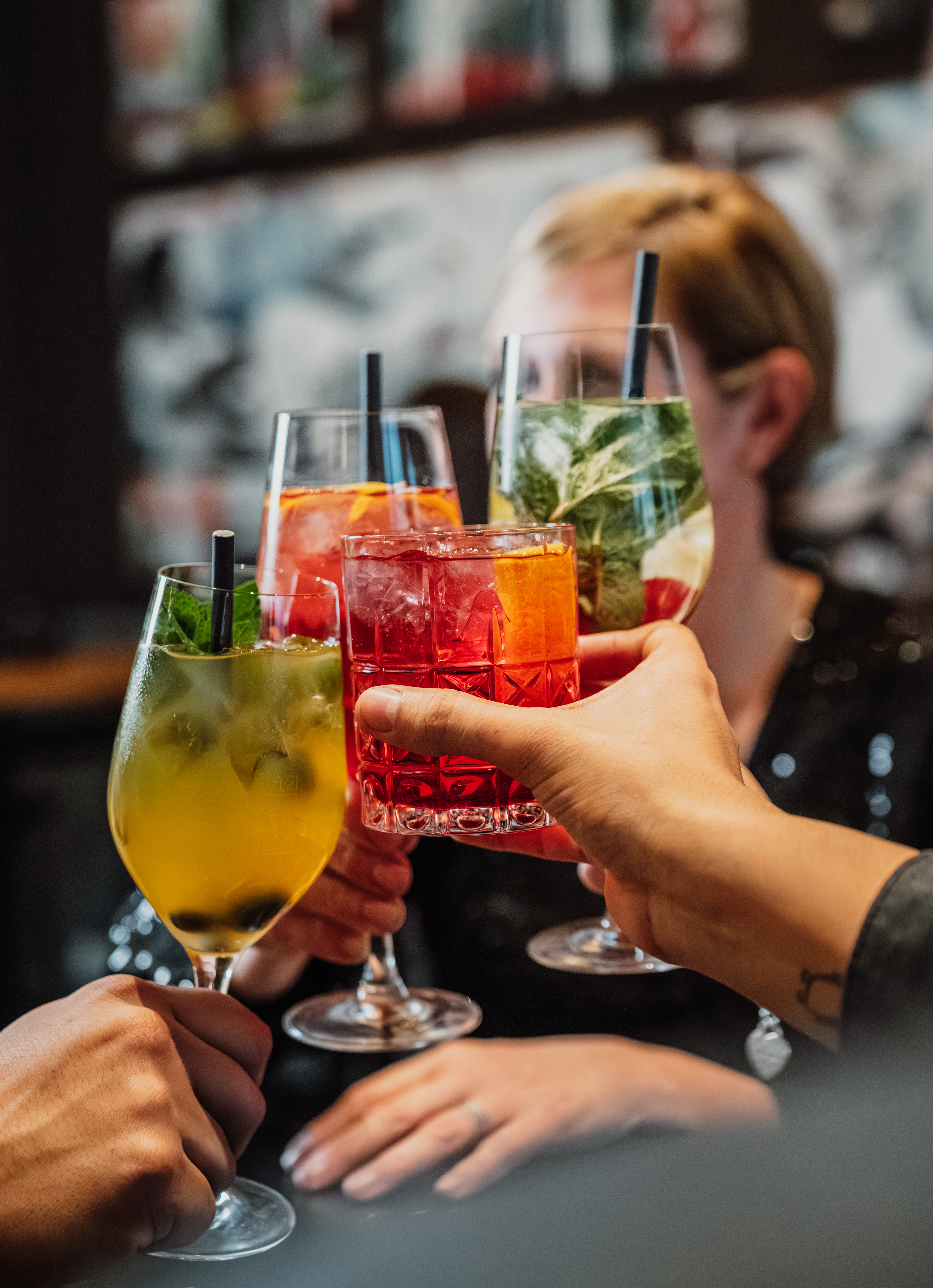 Hands toasting vibrant cocktails with ice, fruit, and mint garnishes.