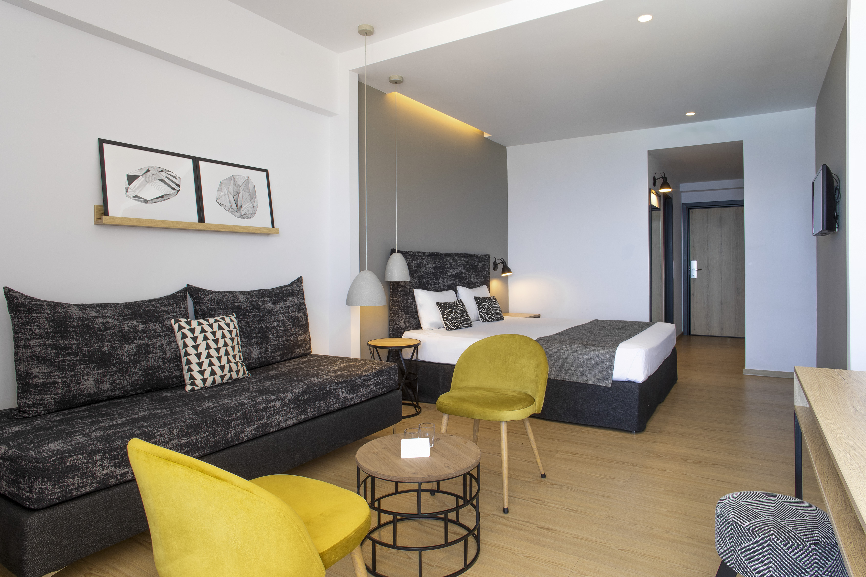 Modern hotel suite with bed, sofa, yellow chairs, geometric art, wood floor.