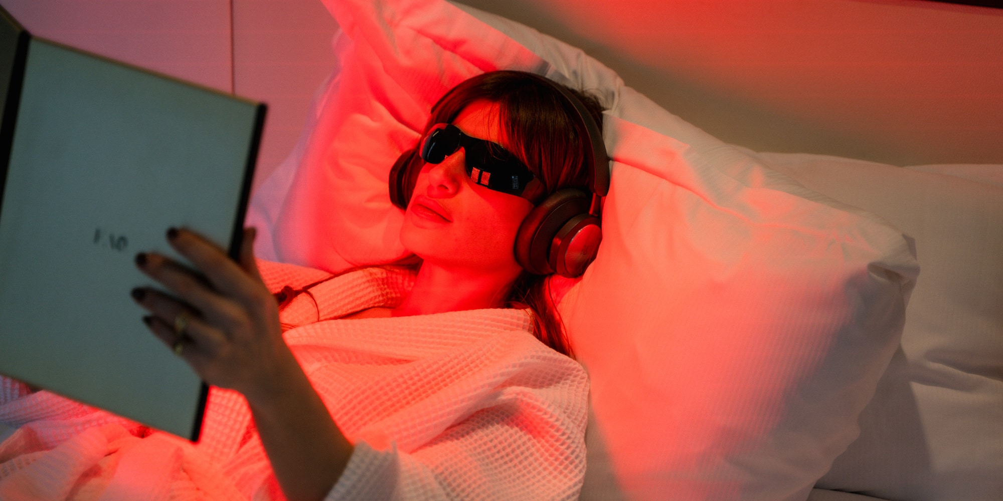 Woman relaxing in bed with red light, therapy glasses, headphones, and book.