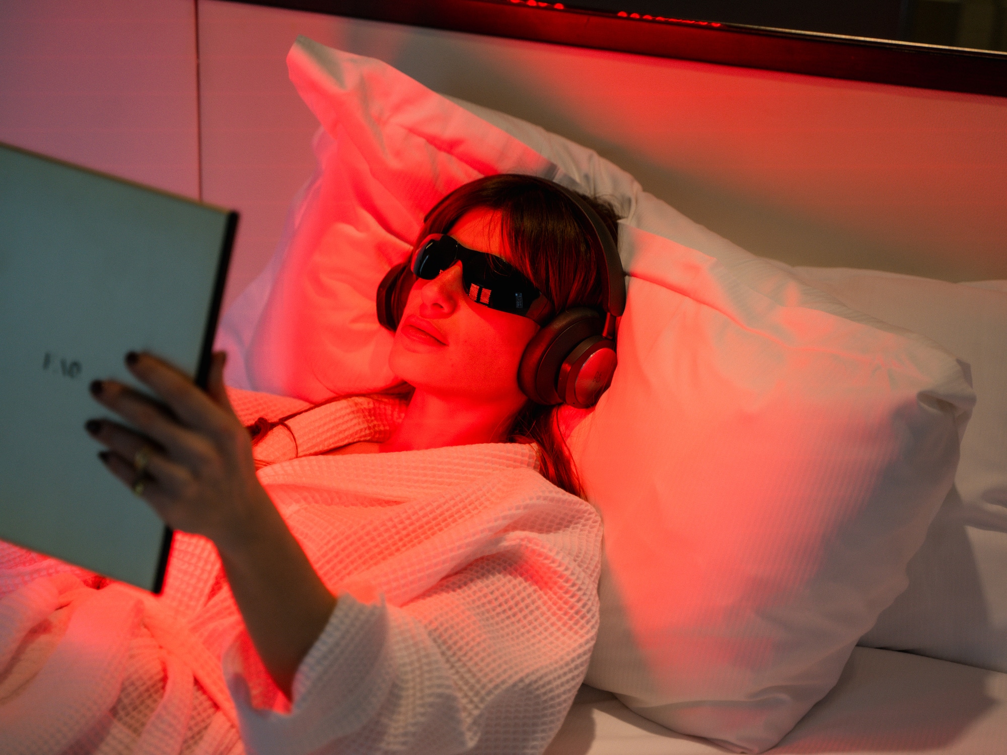 Woman relaxing with red light therapy, eye mask, headphones, and book.