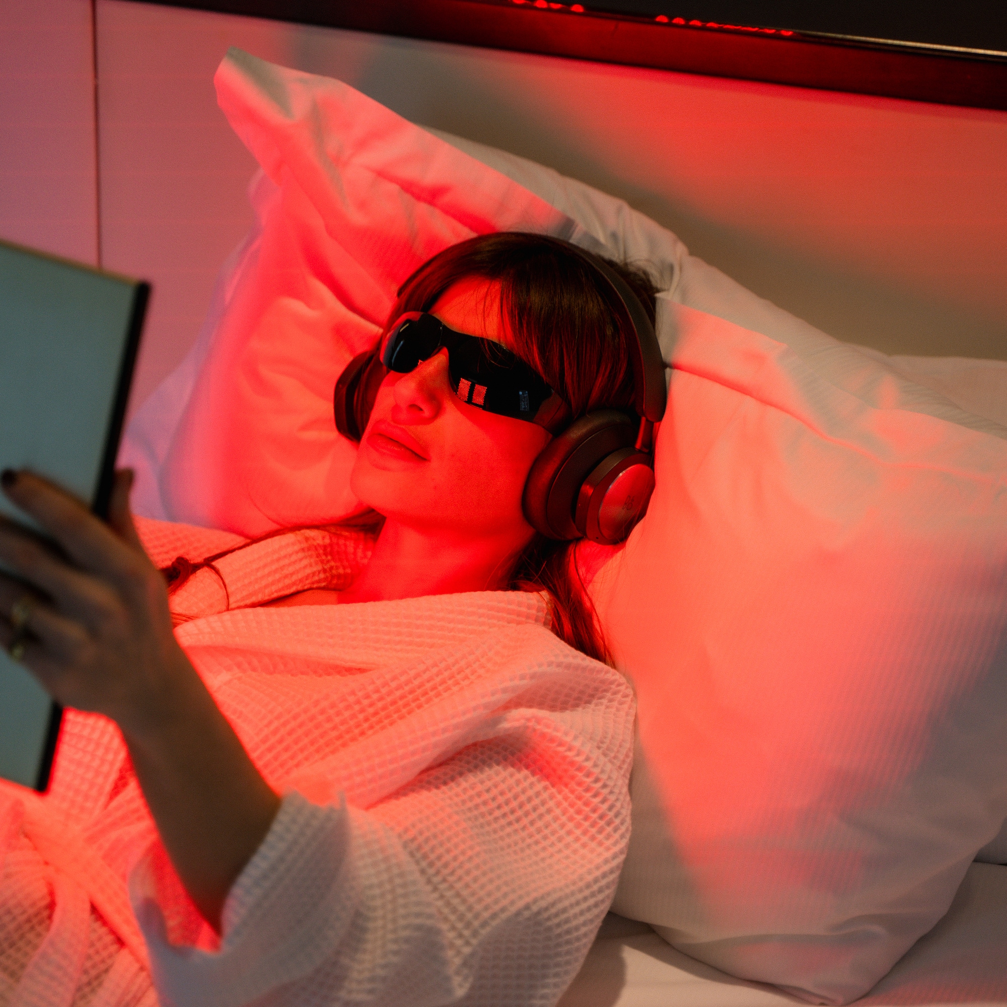 Woman relaxing in bed with red light, therapy glasses, headphones, and book.