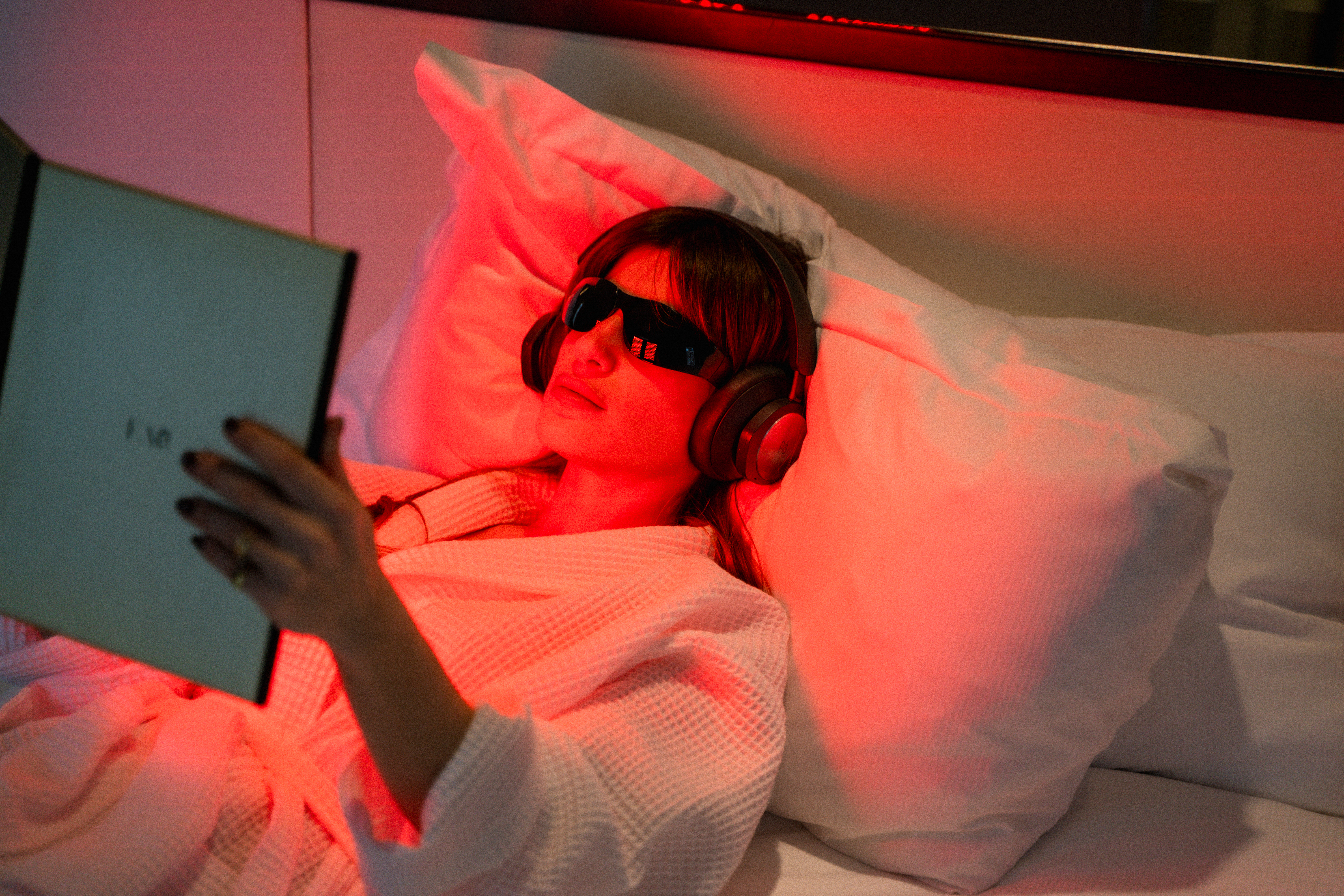 Woman relaxing in bed with red light, therapy glasses, headphones, and book.